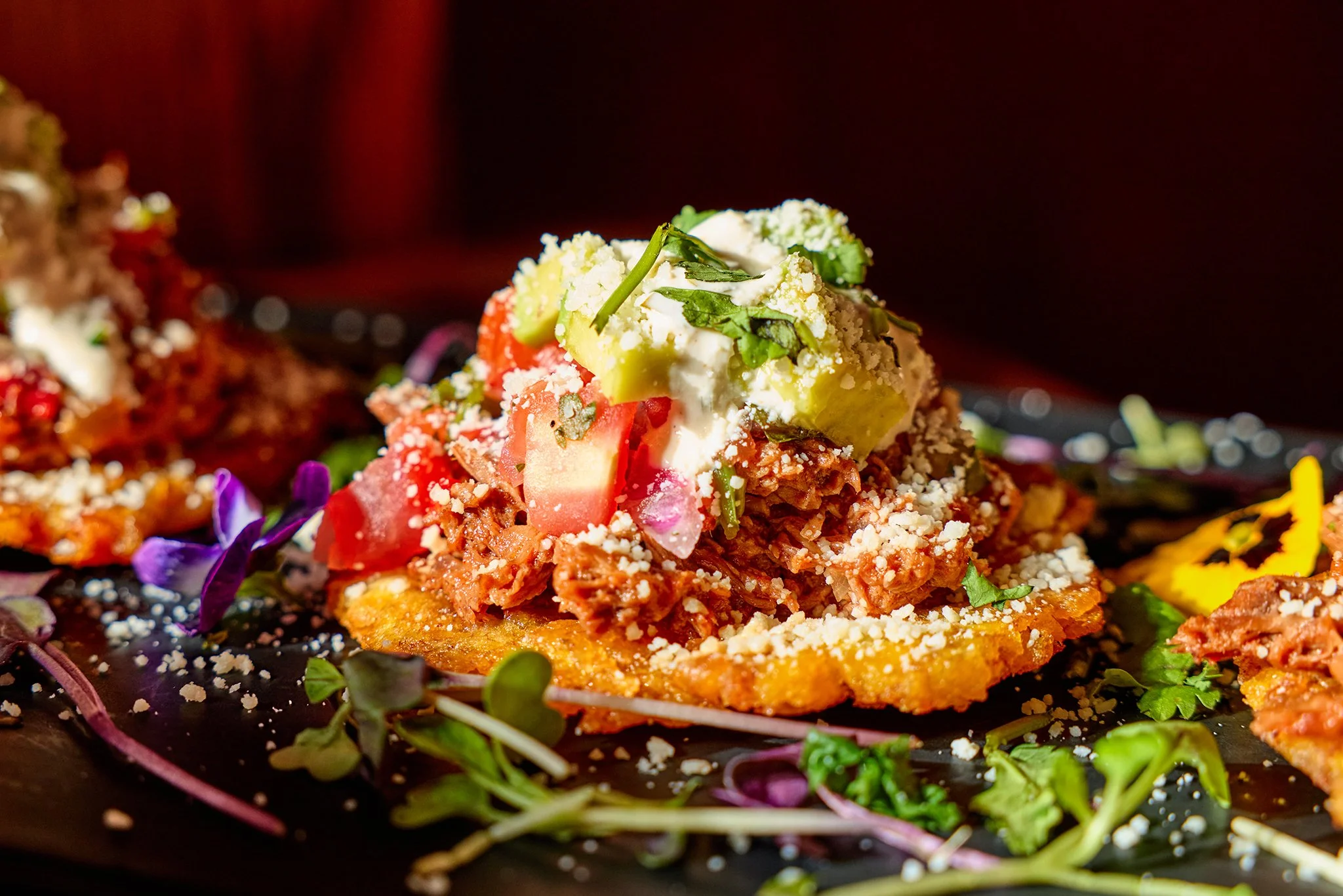 Close-up of a tostada with seasoned ground meat, chopped tomatoes, avocado slices, shredded cheese, and microgreens, on a black plate garnished with edible flowers and herbs.