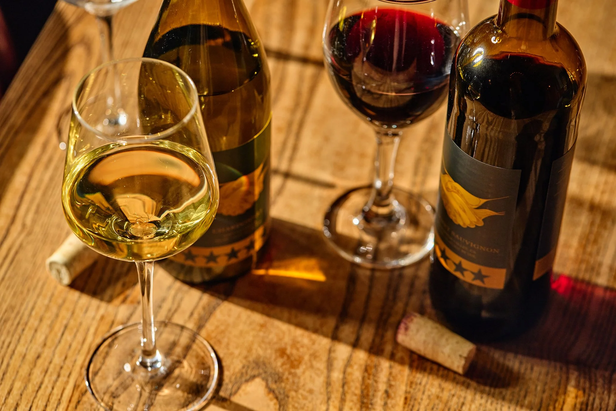 Two bottles of wine, one white and one red, along with a filled white wine glass and an empty red wine glass on a wooden table.