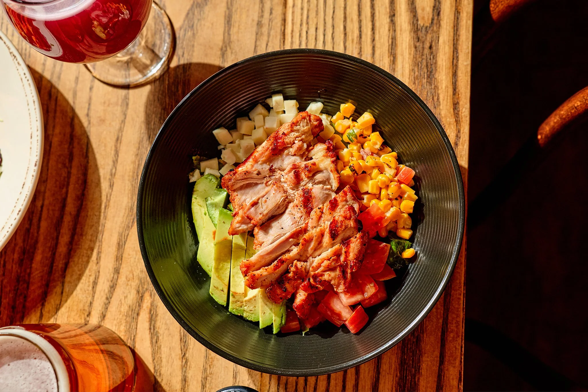 A bowl of chopped vegetables and grilled chicken on a wooden table, with a glass of red wine and a beer nearby.