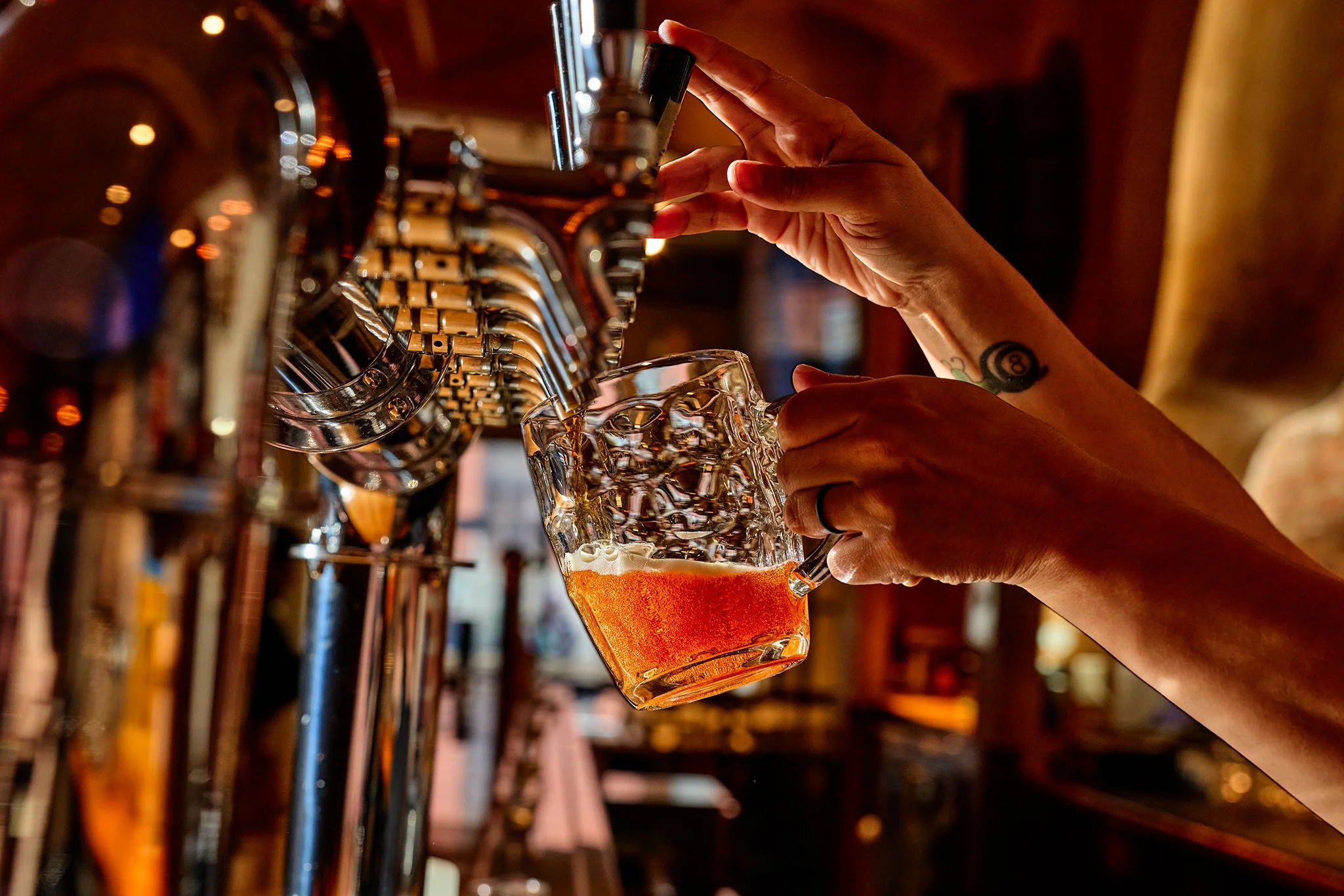 A person pouring a beer from a tap into a glass mug at a bar, with a tattoo on their wrist and a wedding ring on their finger.