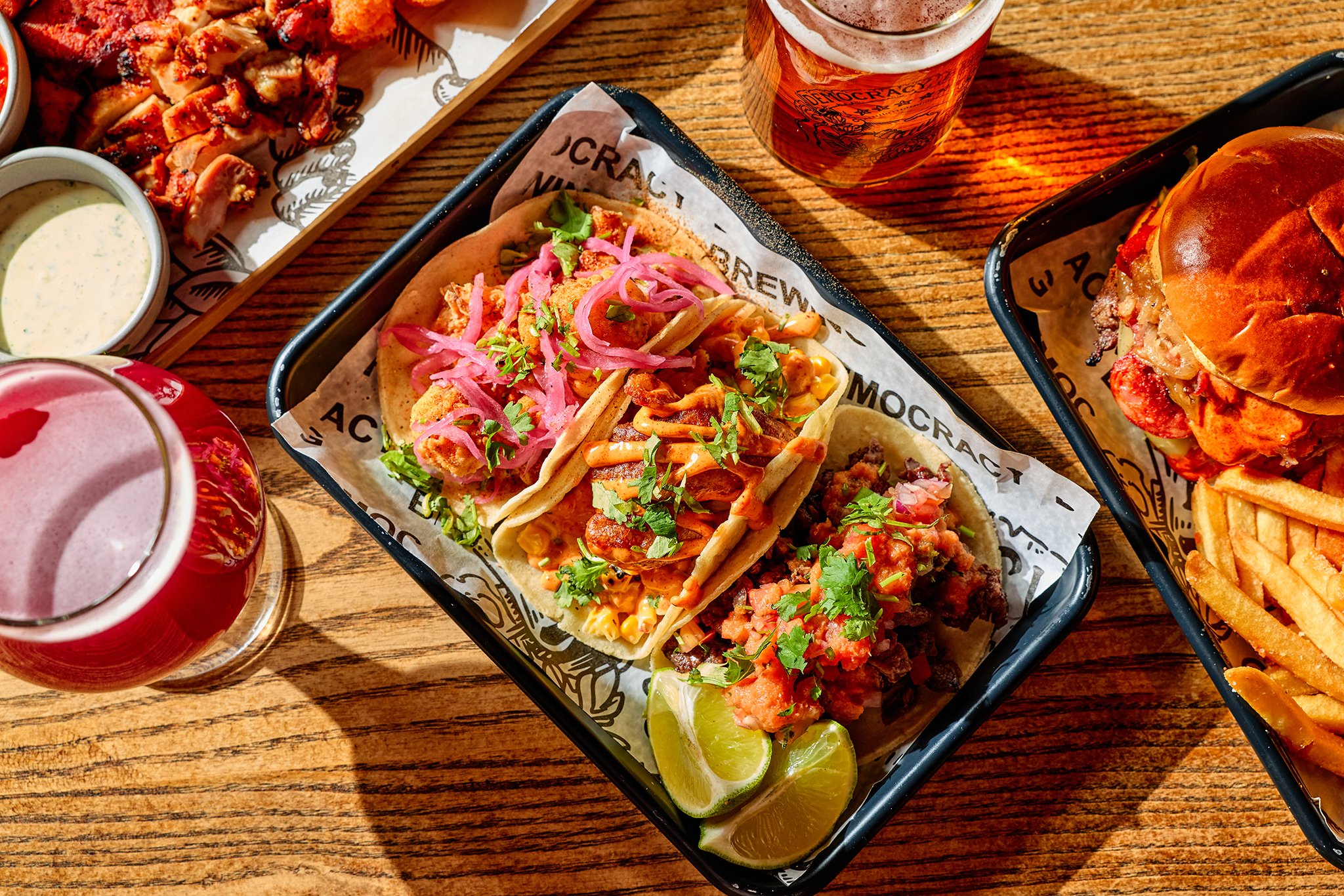 Assorted Mexican dishes on a wooden table, including tacos with pickled onions, sliced lime, a burger with fries, and a small bowl of sauce.