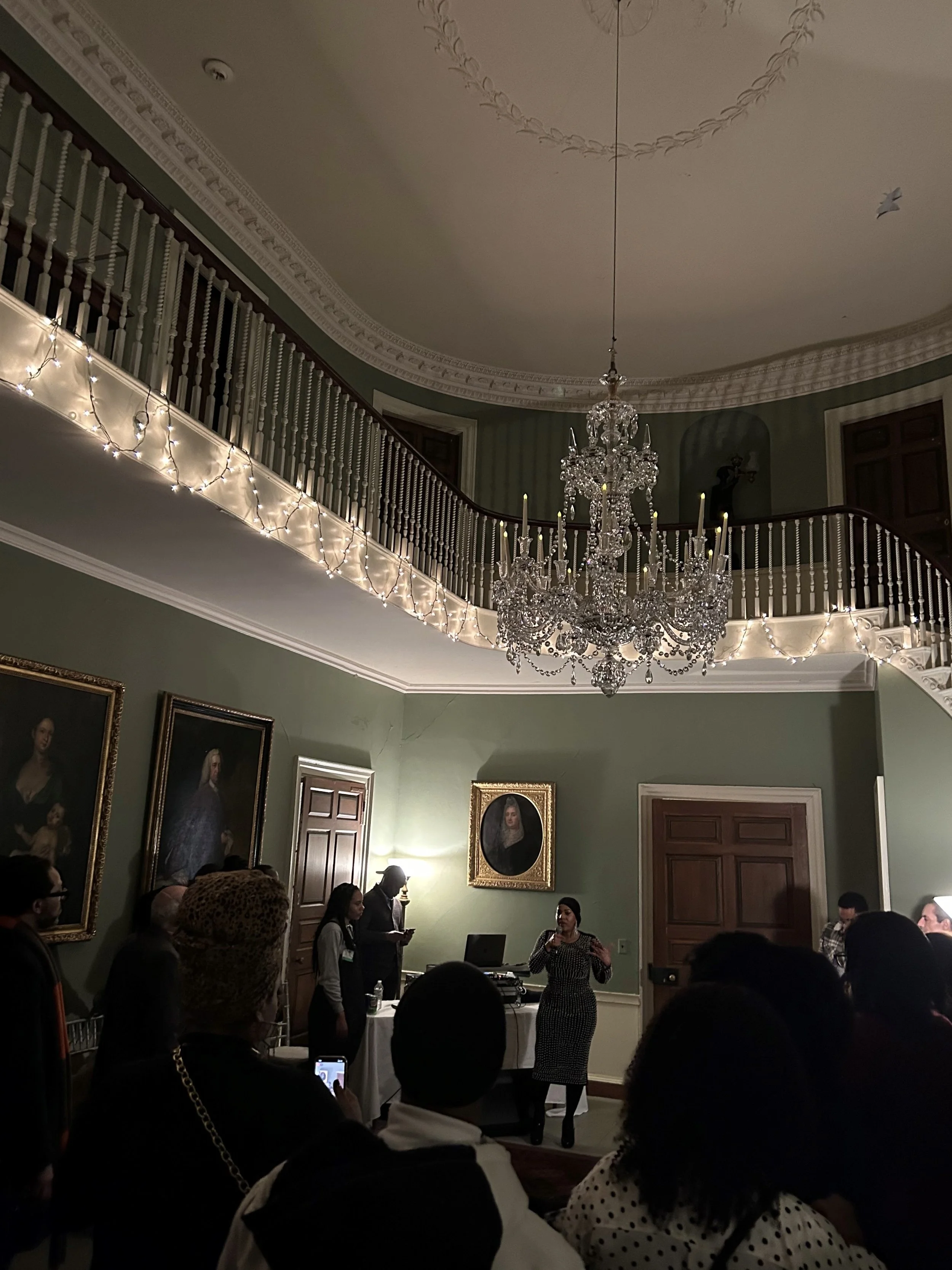 People attending a presentation or speech in an elegant, historic room with a chandelier, paintings, and string lights on the balcony.