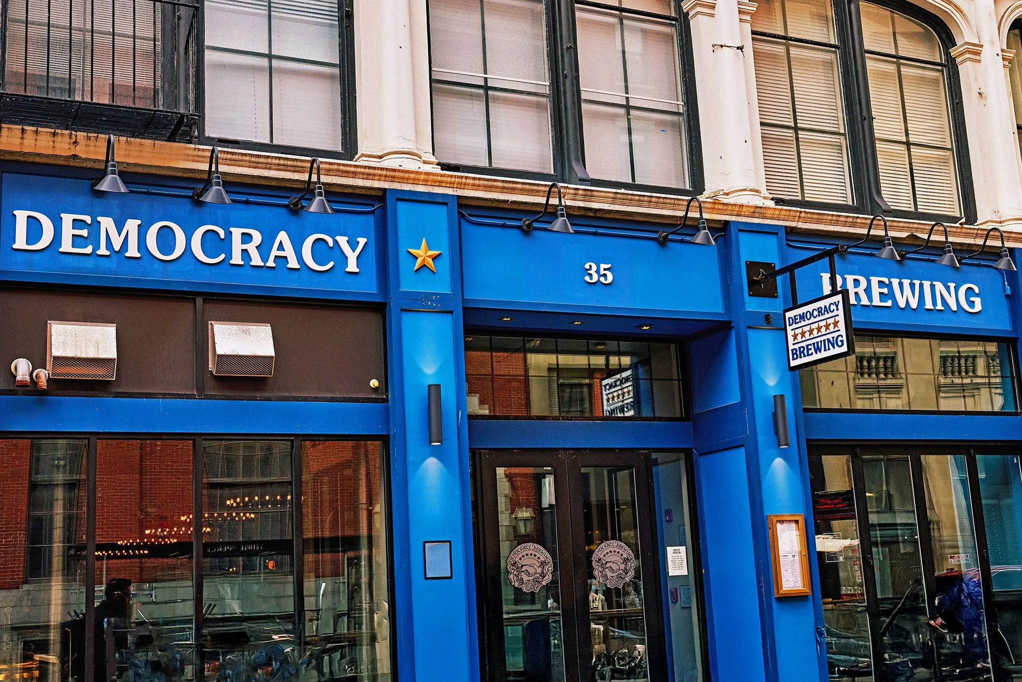 Exterior of a building with blue signage reading 'Democracy Brewing' and the number 35, with large windows on upper floors and glass doors on the ground floor.