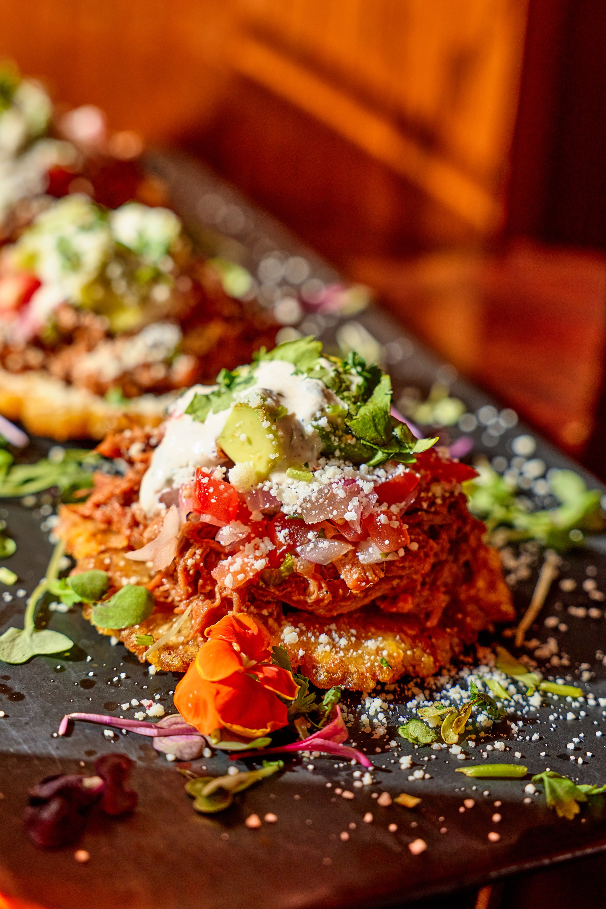 Close-up of two traditional Mexican tacos topped with diced tomatoes, onions, avocado, sour cream, and cilantro, garnished with edible flowers and microgreens on a black slate plate.