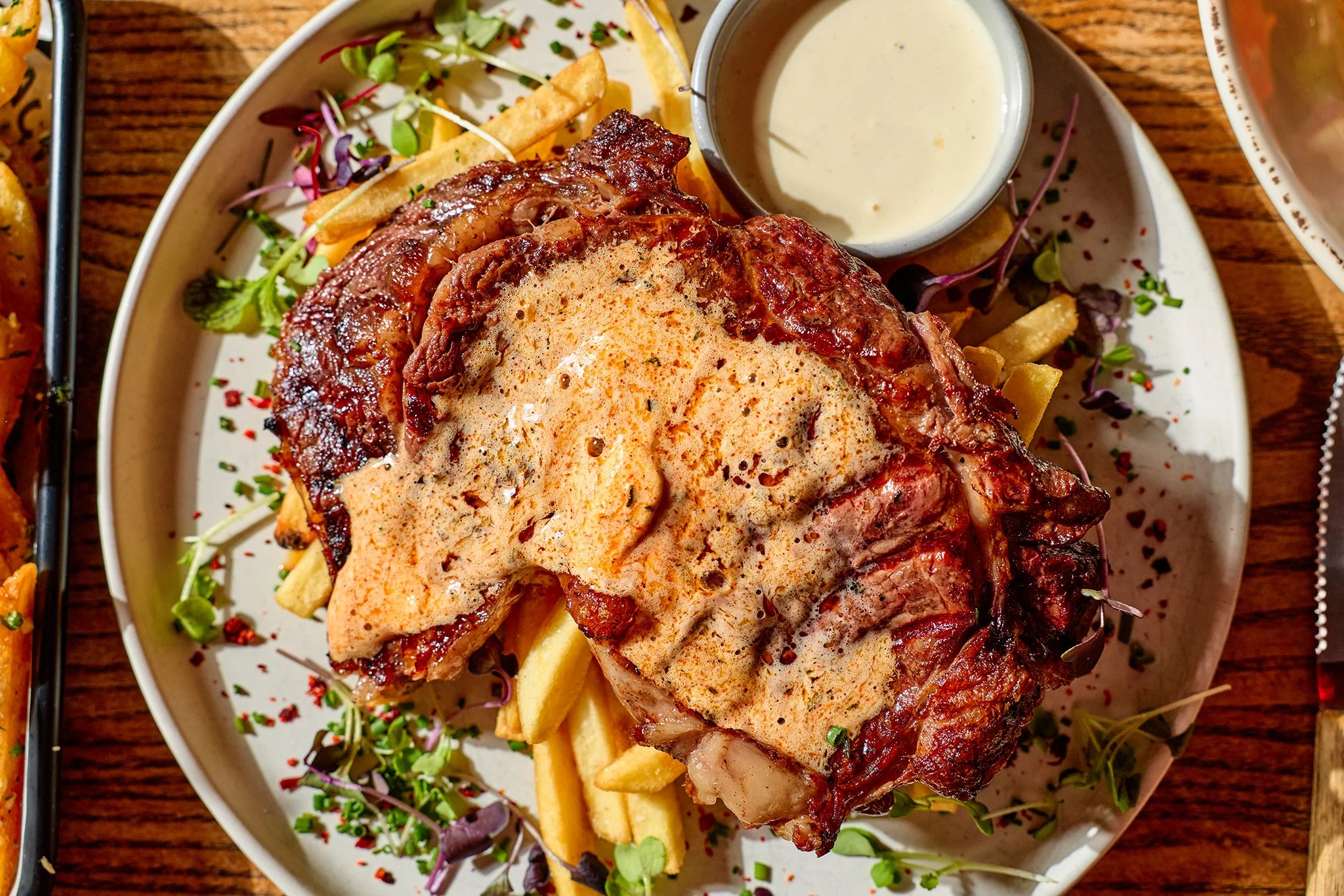 Steak topped with a creamy sauce, served with French fries, microgreens, and a side of creamy white dipping sauce.