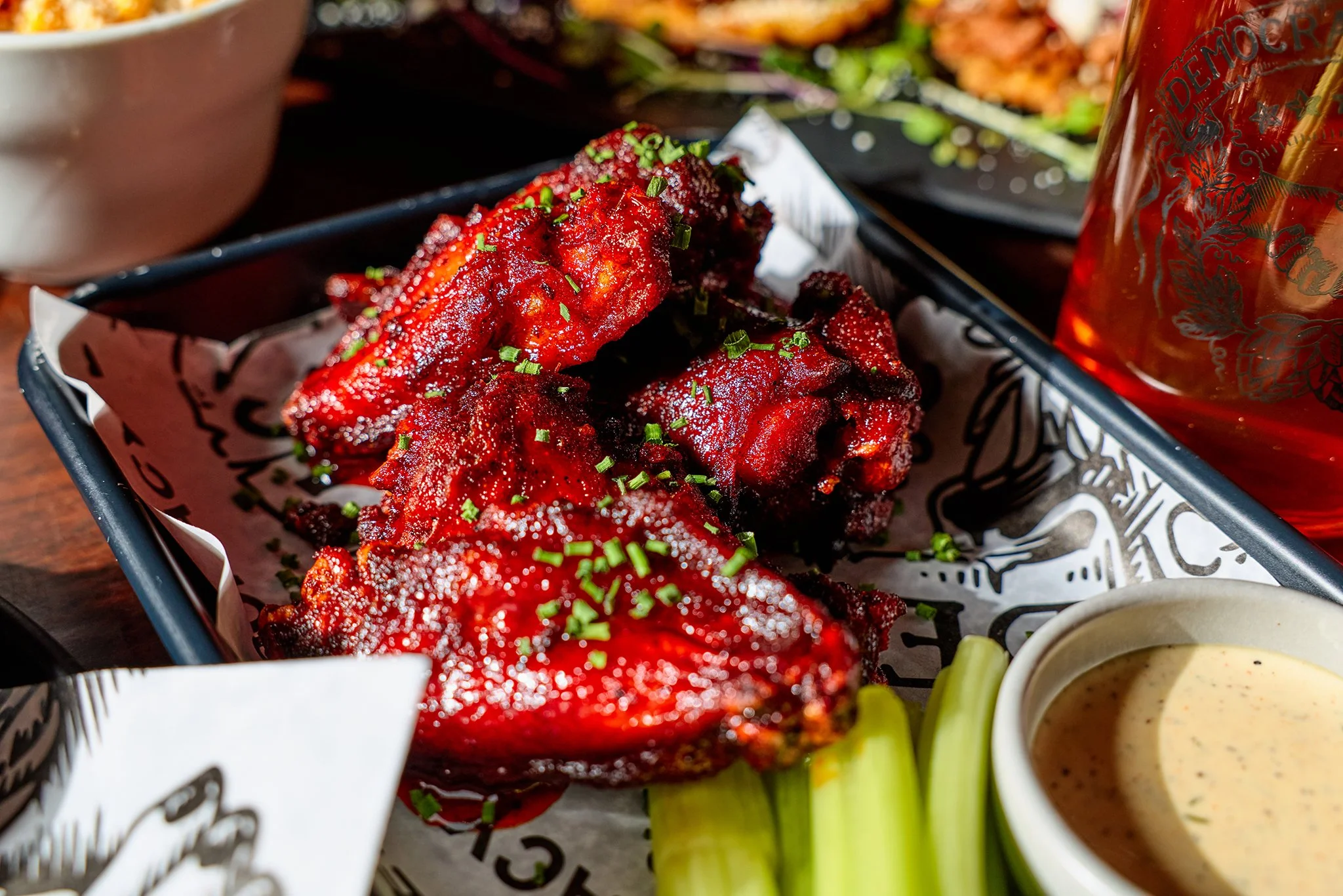 Buffalo chicken wings with chopped green onions, served with a side of creamy dipping sauce and celery sticks, on a black tray lined with printed parchment paper, with a glass of beer nearby.