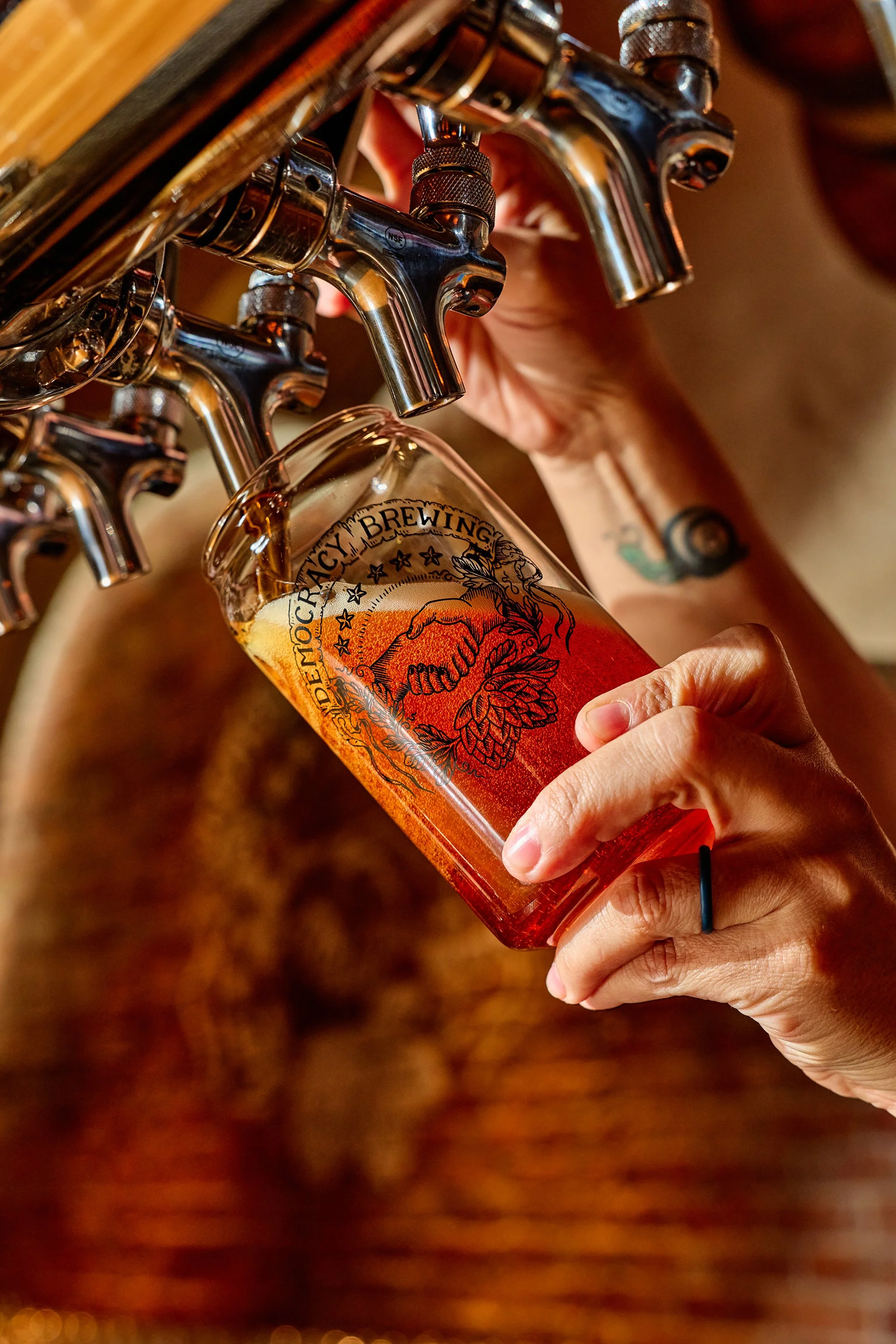 A person pours amber-colored beer from a tap into a pint glass with a logo that says 'Defiance OCC' and features a handshake within a decorative crest.