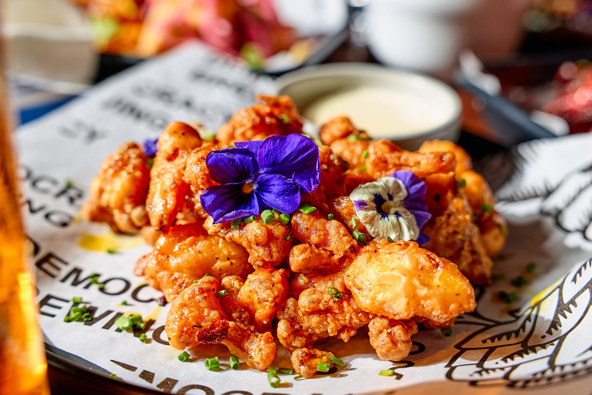 Fried chicken topped with purple and white edible flowers, garnished with chopped green herbs, served with a side of dipping sauce.