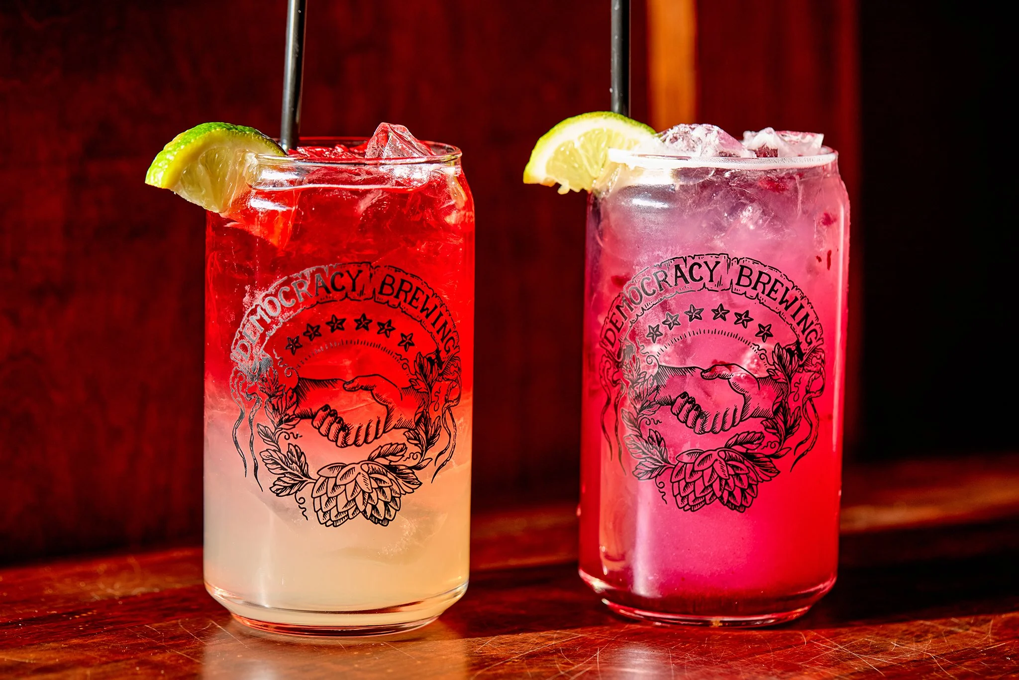 Two colorful cocktails in tall glasses with 'Democracy Brewing' logos, garnished with lemon wedges, on a wooden surface.
