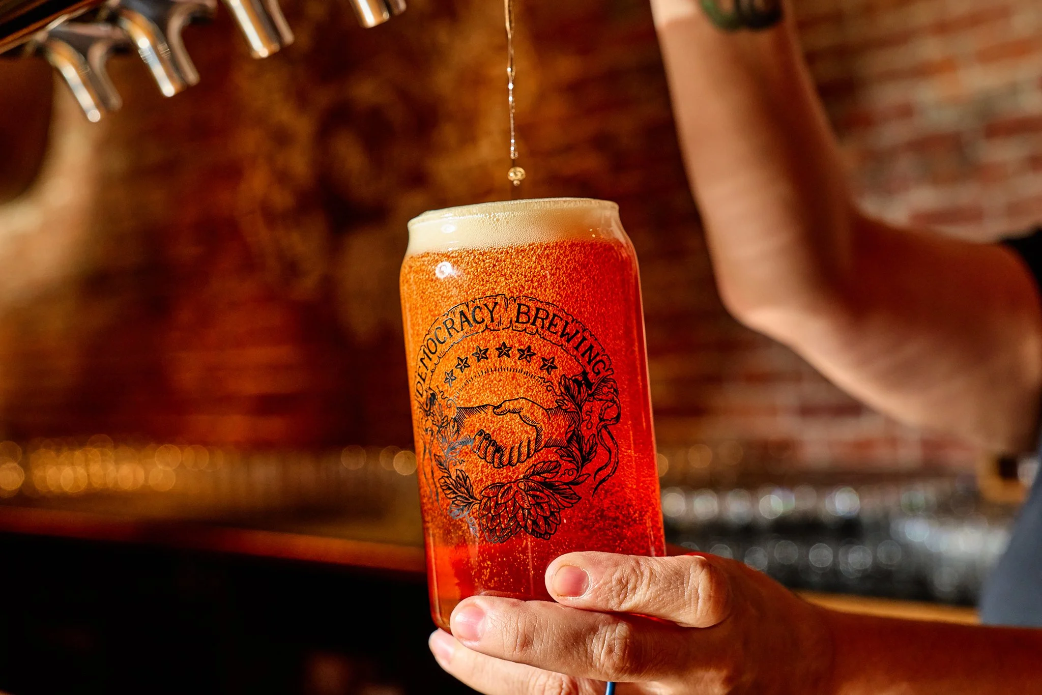 A person holds a tall glass of amber-colored beer with a foamy head, with a droplet falling from the tap at a brewery or bar.