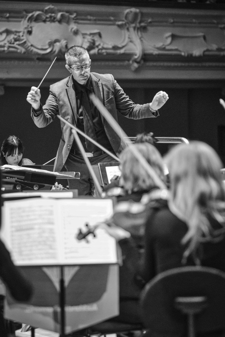 A conductor leading an orchestra during a performance, with musicians reading sheet music in front of them.
