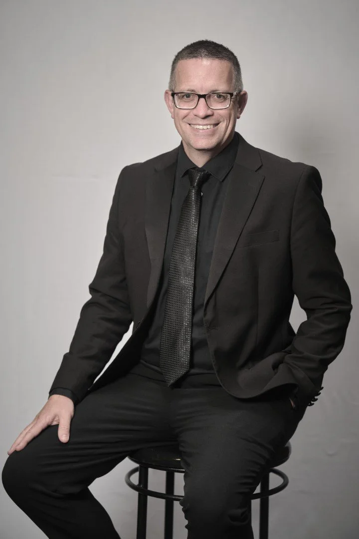 A man in a black suit, black shirt, and black tie sitting on a stool, smiling, with a light gray background.