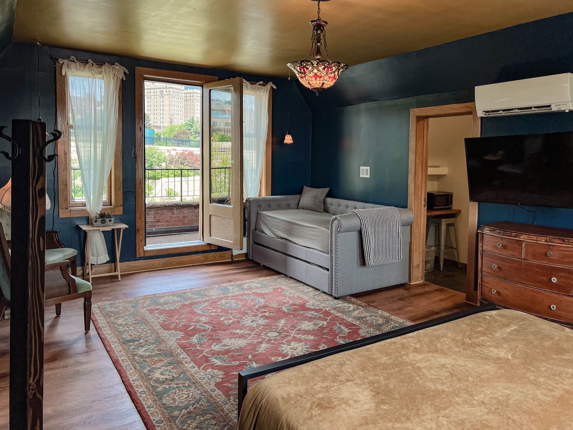 Living room with large windows and a balcony door, a gray daybed with a pillow and striped blanket, a wooden dresser with a flat-screen TV mounted above, an air conditioning unit, an ornate ceiling lamp, and a colorful area rug on wooden floors.