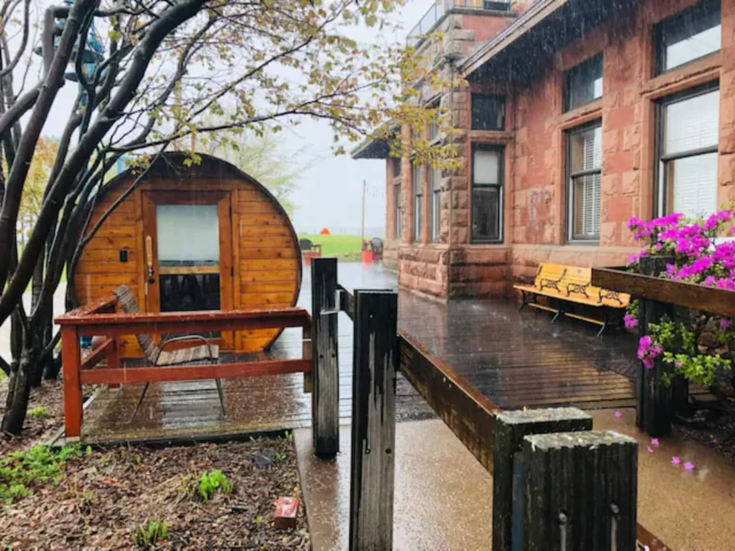 Rainy outdoor scene showing a small wooden sauna, a bench, and a patio with purple flowers on a flower box, next to a red brick building with multiple windows and picnic tables in the distance.