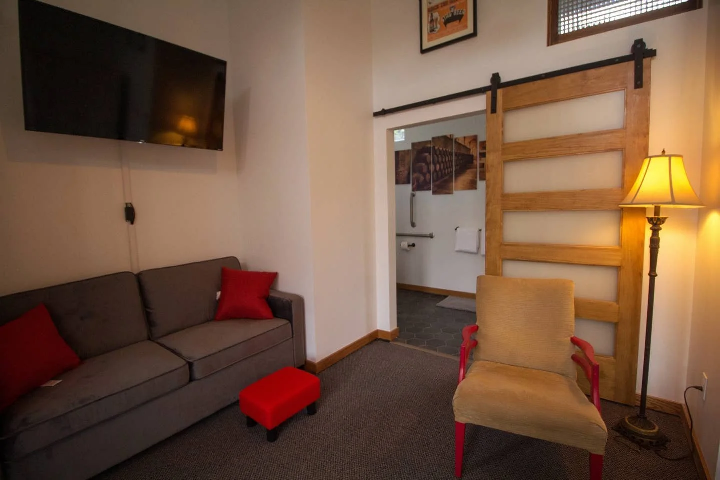 Living room with a wall-mounted flat-screen TV, a sofa with red pillows, a wooden sliding barn door, a beige armchair with red arms, and a tall floor lamp illuminating the room.