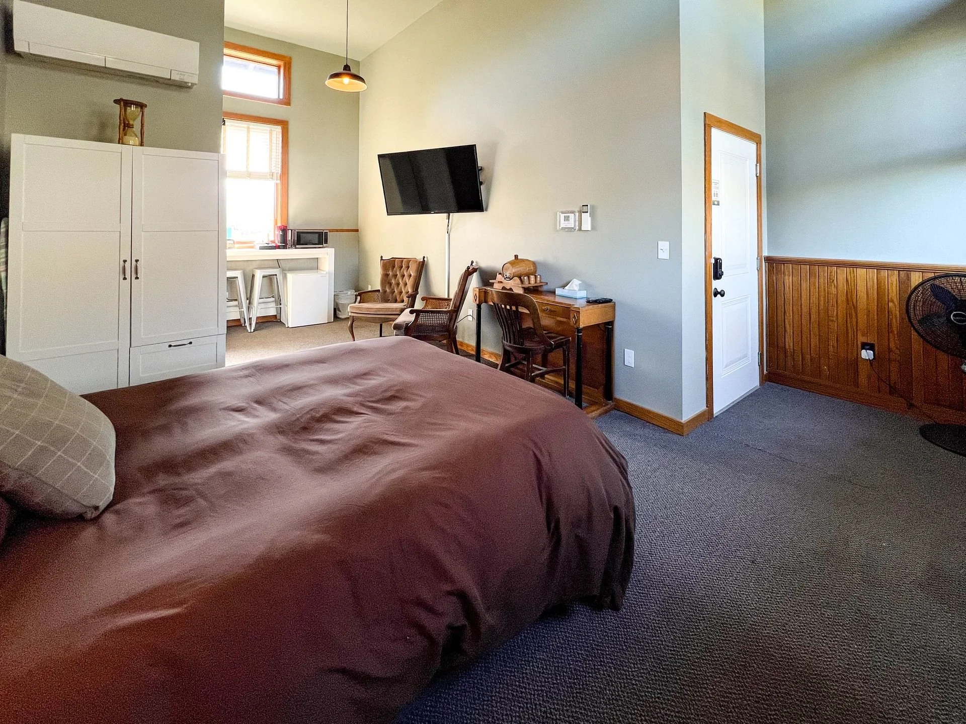 A cozy hotel room with a bed, a wall-mounted TV, a small workspace, a white wardrobe, and a sitting area near windows with red frames, wooden accents, and neutral-colored walls.
