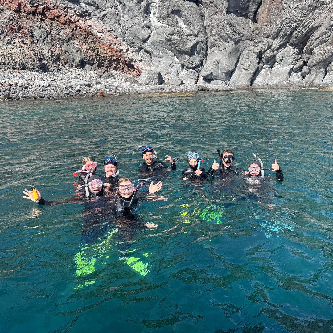 Group of people wearing snorkel masks and wetsuits floating together in clear water near a rocky shoreline