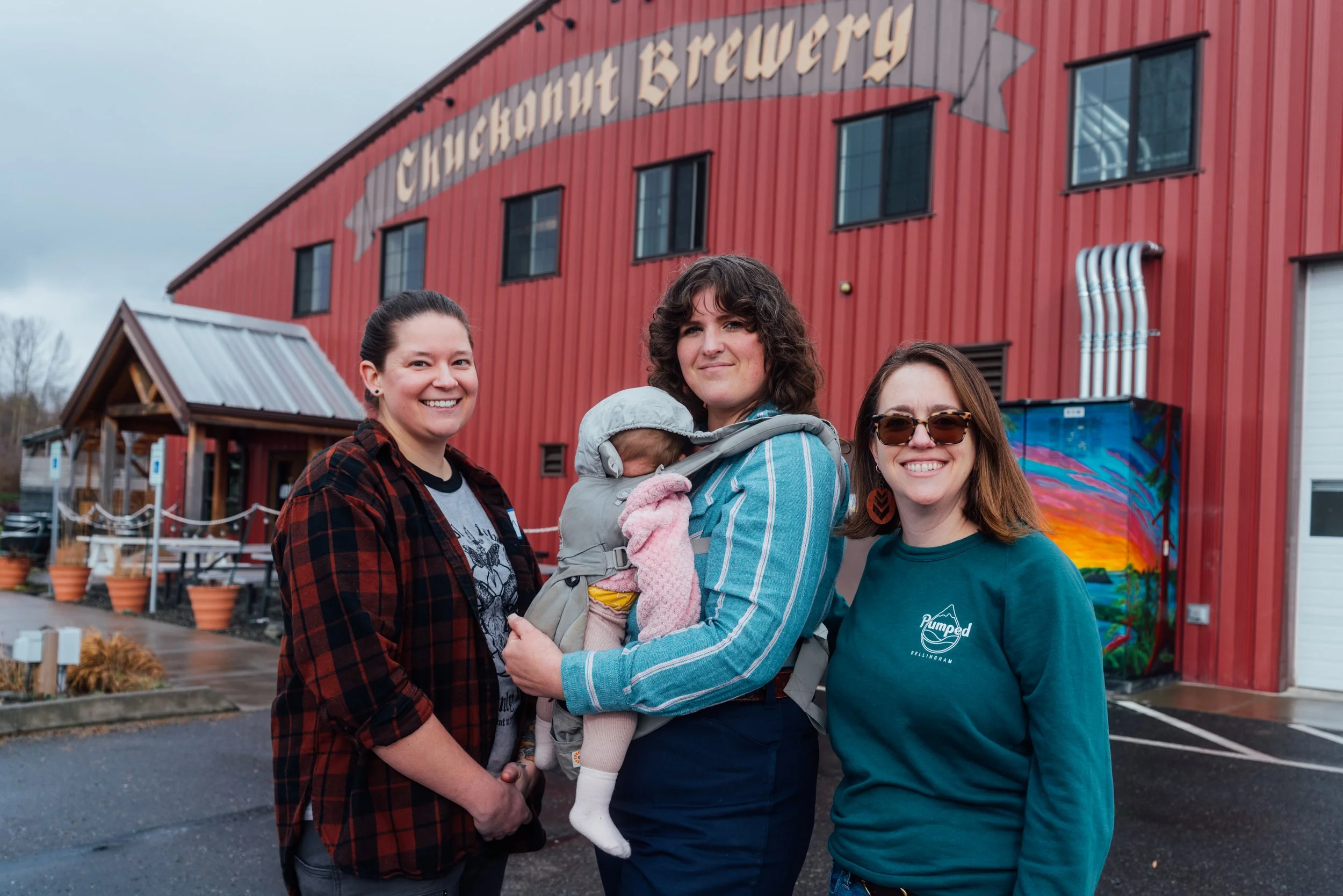 Group of four women, one holding a baby in a gray carrier, standing outside Chickahout Brewery in front of a red building with a colorful mural.