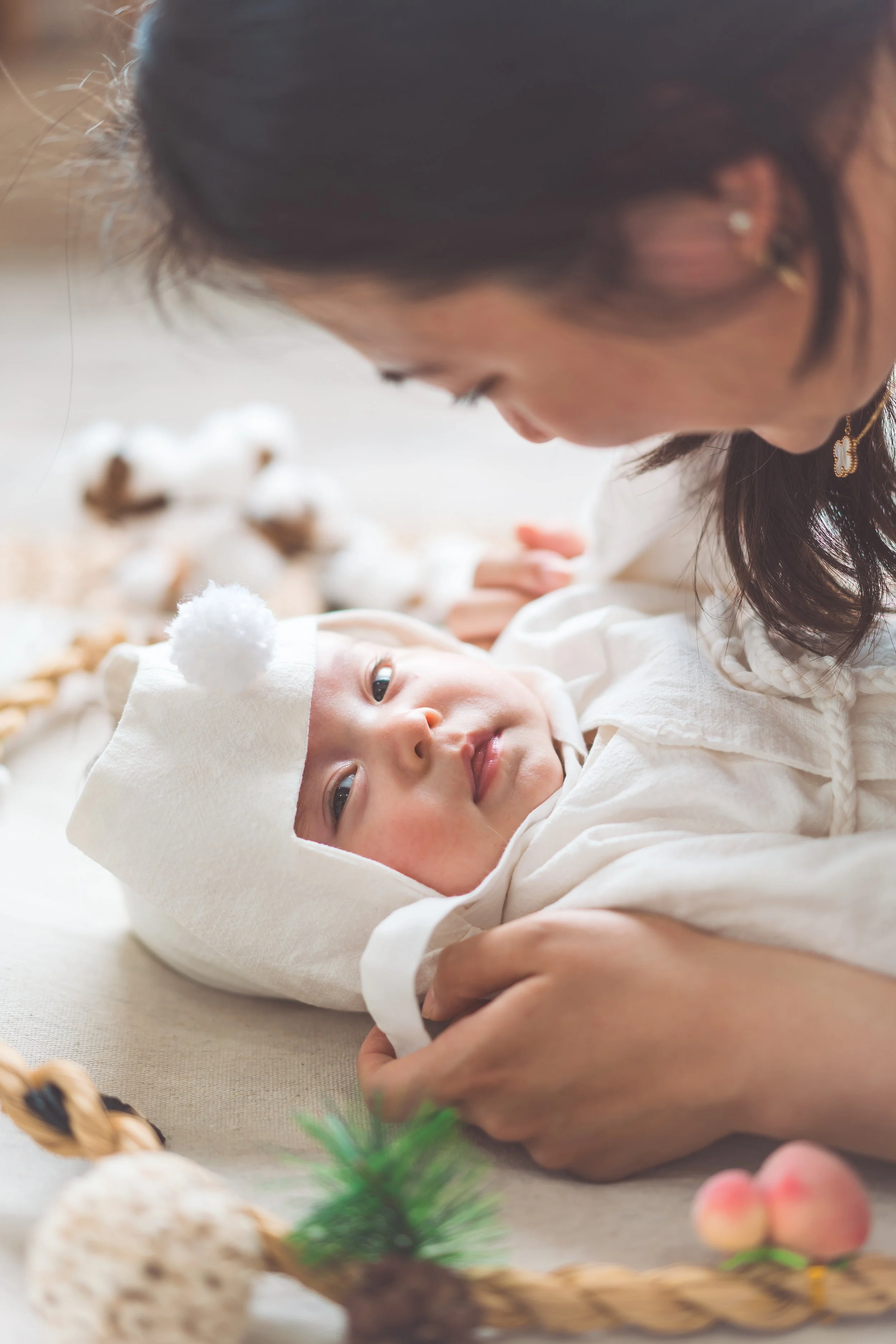 Woman lying next to a baby dressed in white with a white hat, looking at him lovingly amidst Christmas decorations.
