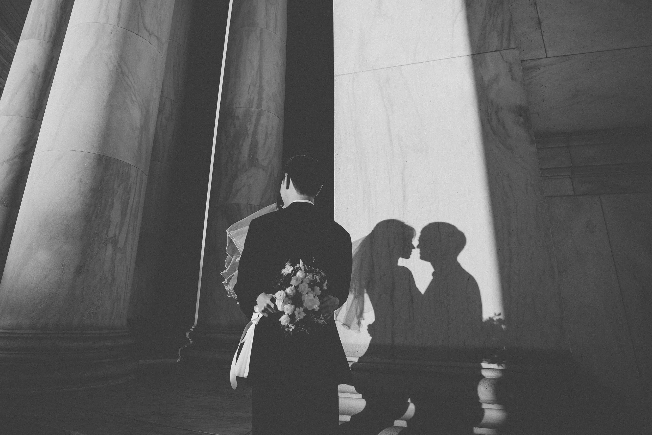 A bride and groom, with their backs to the camera, share a moment as their shadows are cast on a marble wall, inside a building with large marble columns.