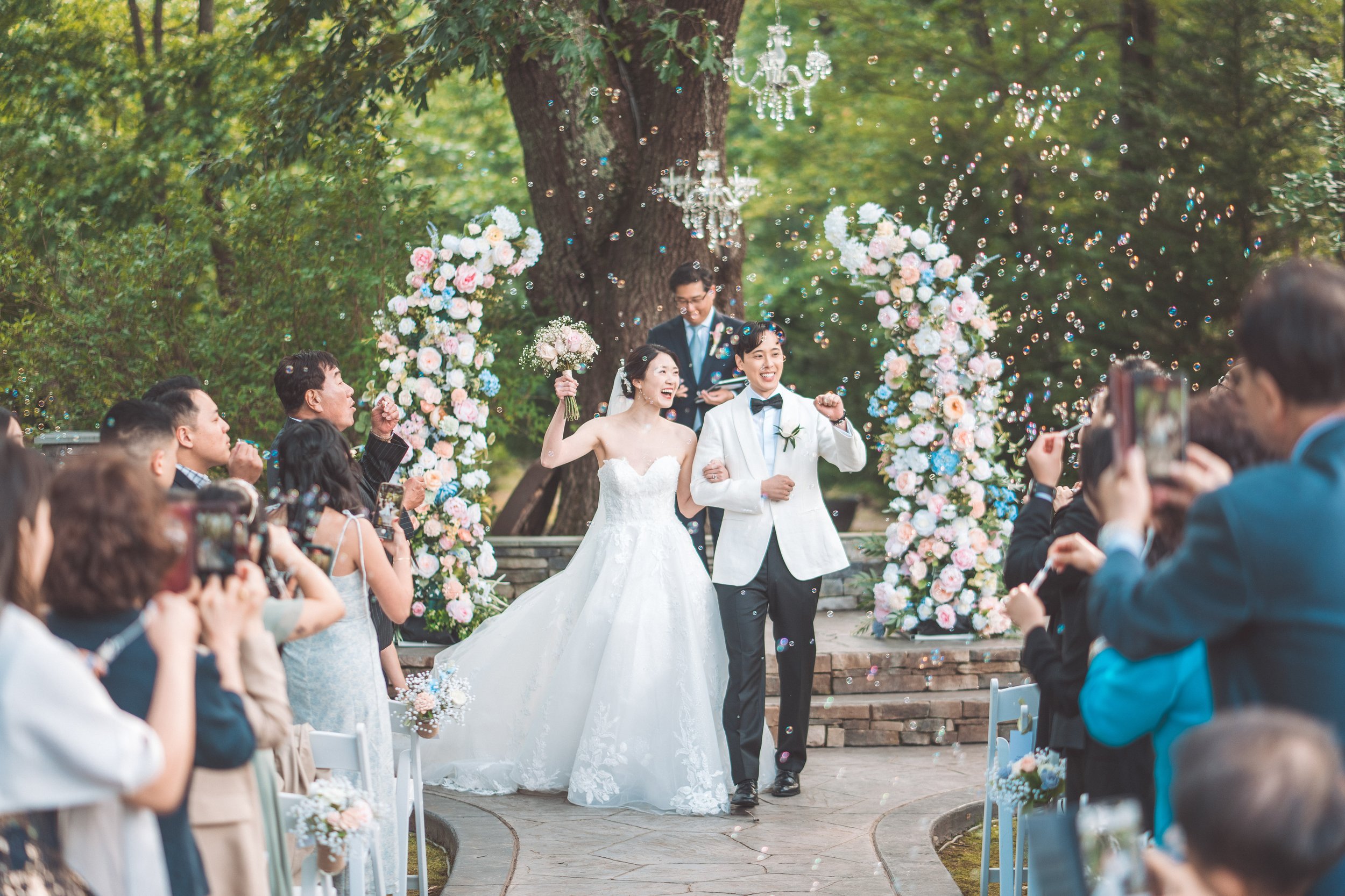 A newlywed couple walks down the aisle at an outdoor wedding ceremony, celebrating with guests, surrounded by floral decorations and a large tree in the background.