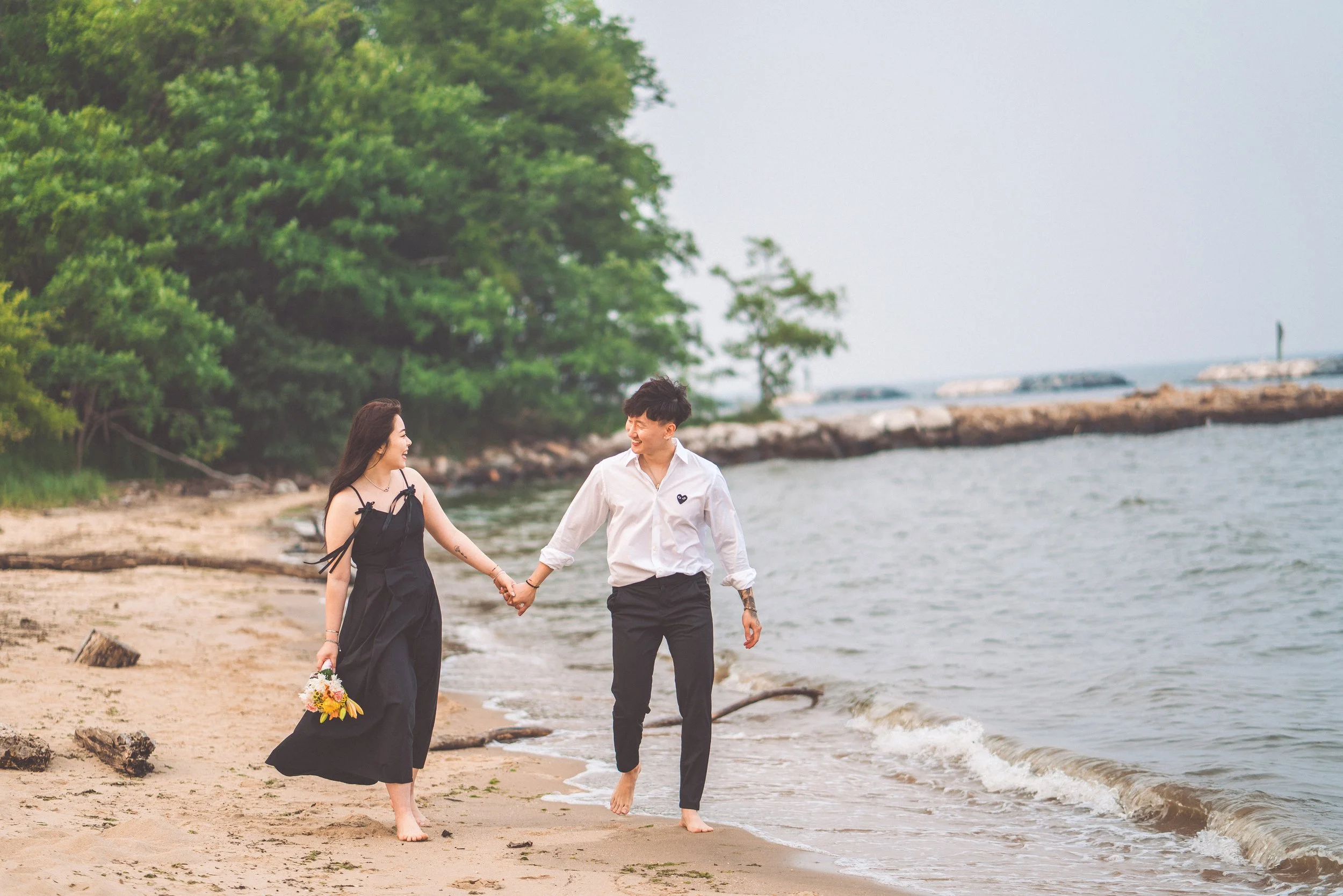 A couple walking hand in hand along the beach with trees in the background, smiling and holding a bouquet of flowers.