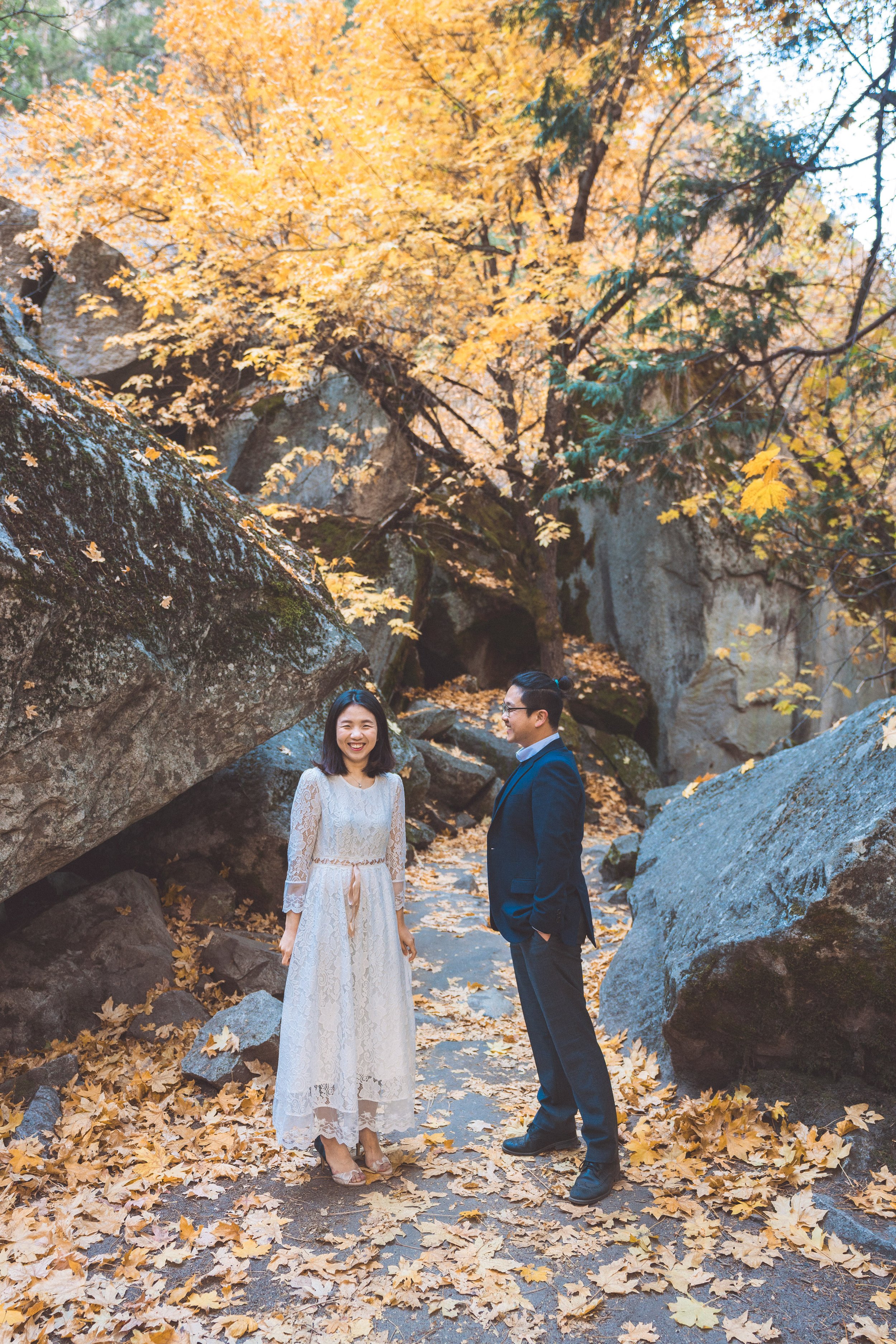 A woman in a white lace dress and a man in a navy suit standing outdoors on a fall day with yellow leaves on the ground and trees in the background.