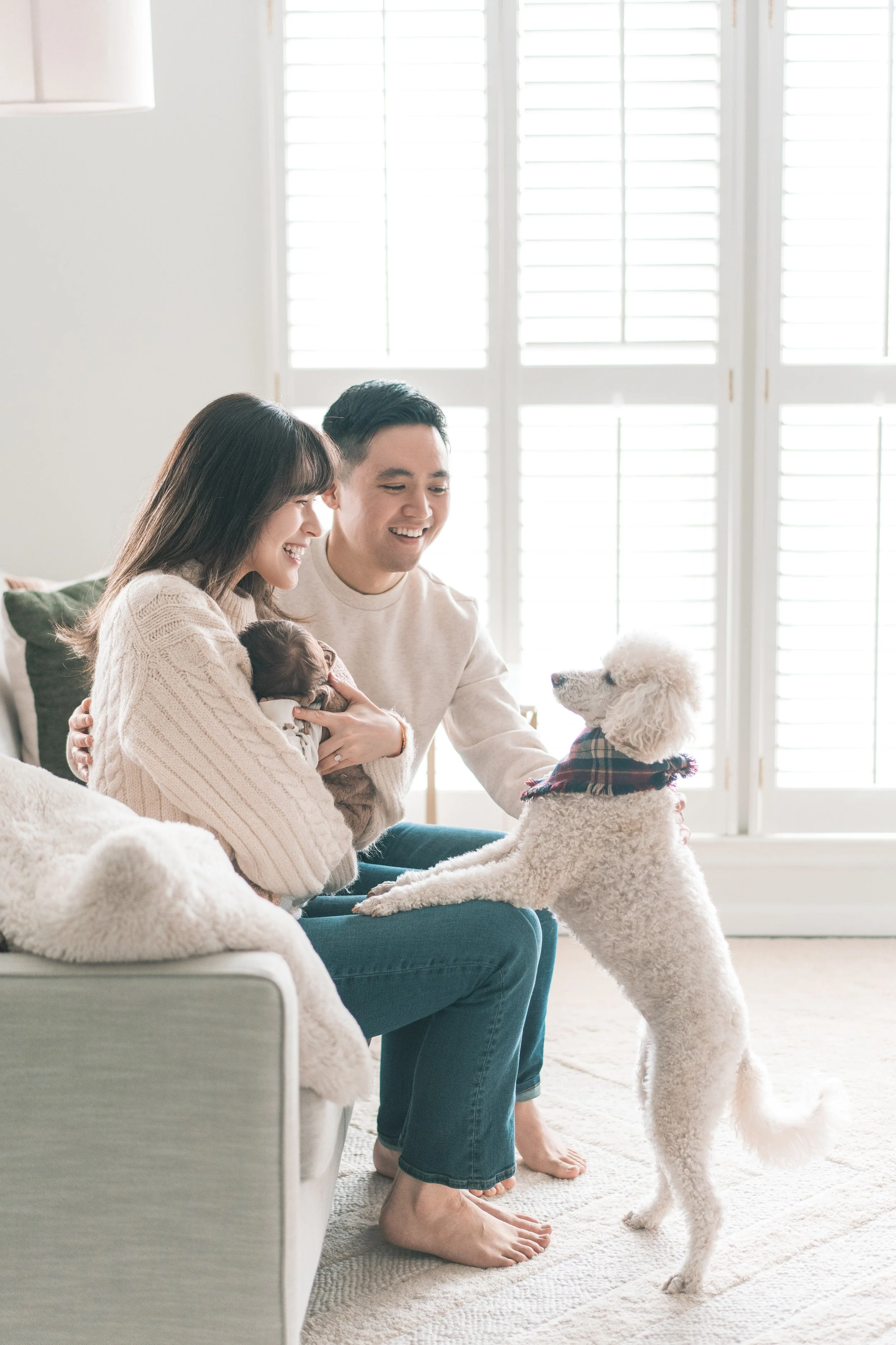 A smiling couple sitting on a sofa with their small baby and a white poodle dog wearing a bandana, in a bright room with large windows.