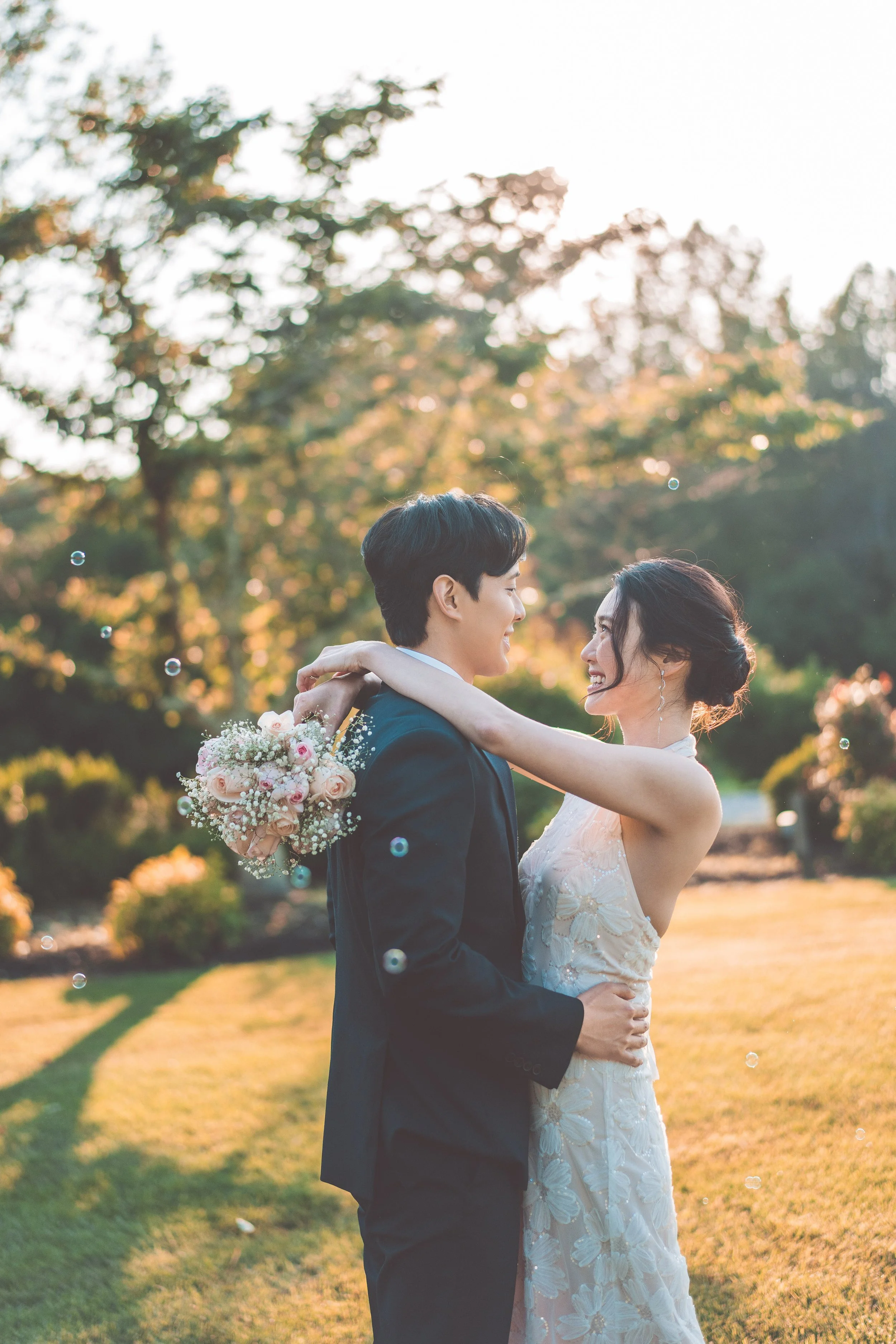 A bride and groom embrace outdoors in a park during sunset, with the bride holding a bouquet of roses and baby's breath.