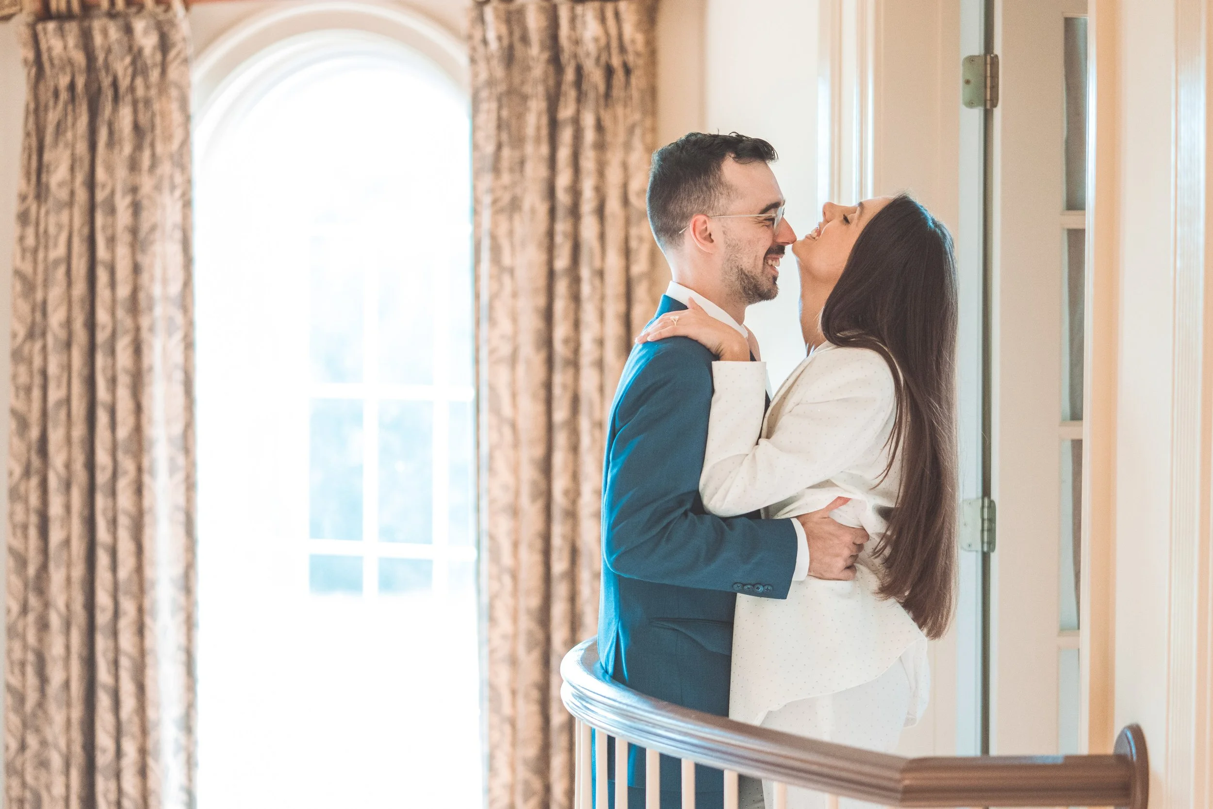 A couple in wedding attire sharing an intimate moment indoors near a window with curtains.