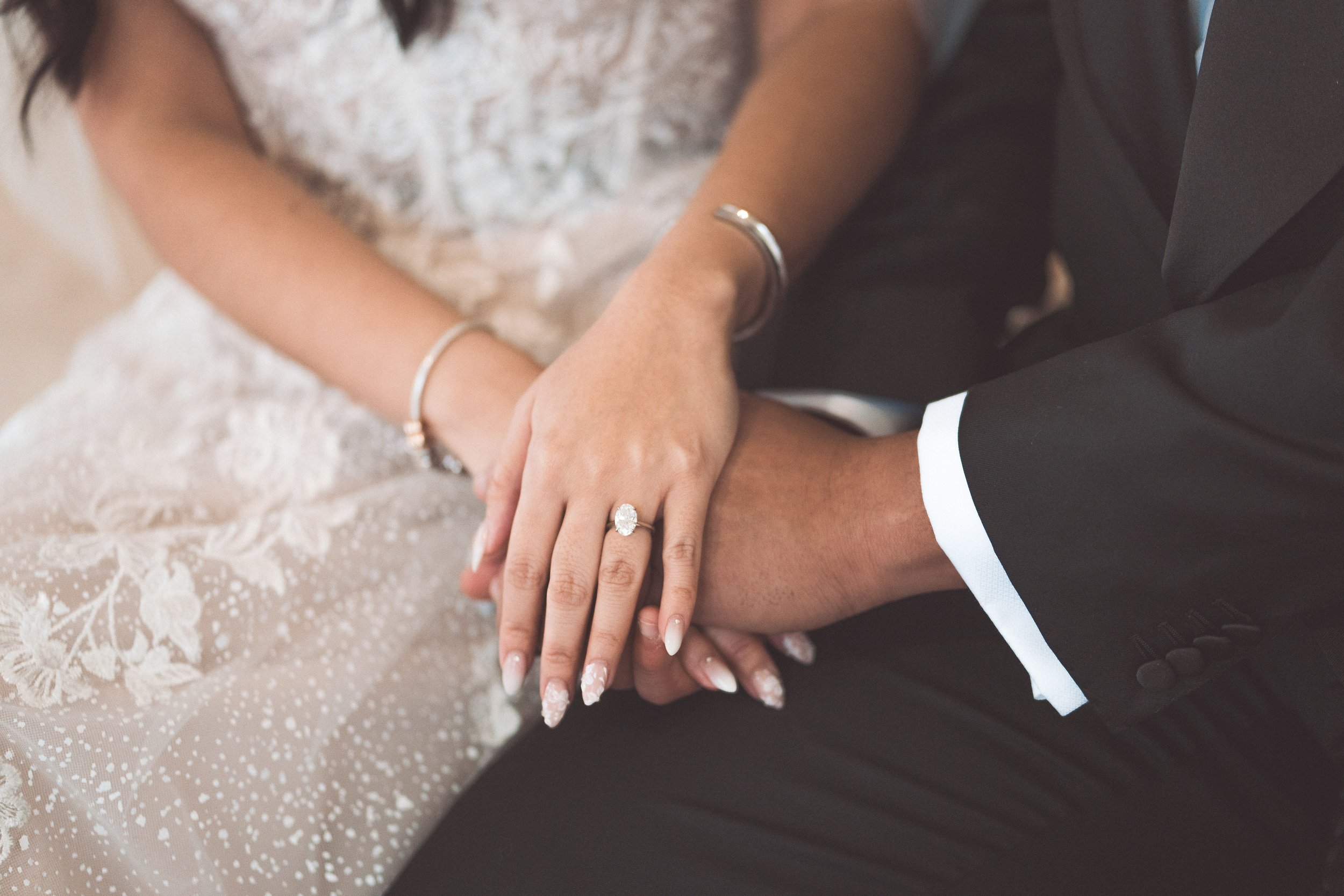 Close-up of a woman and man holding hands, showing an engagement ring and wedding bands, with woman's lace wedding dress and man's black suit.