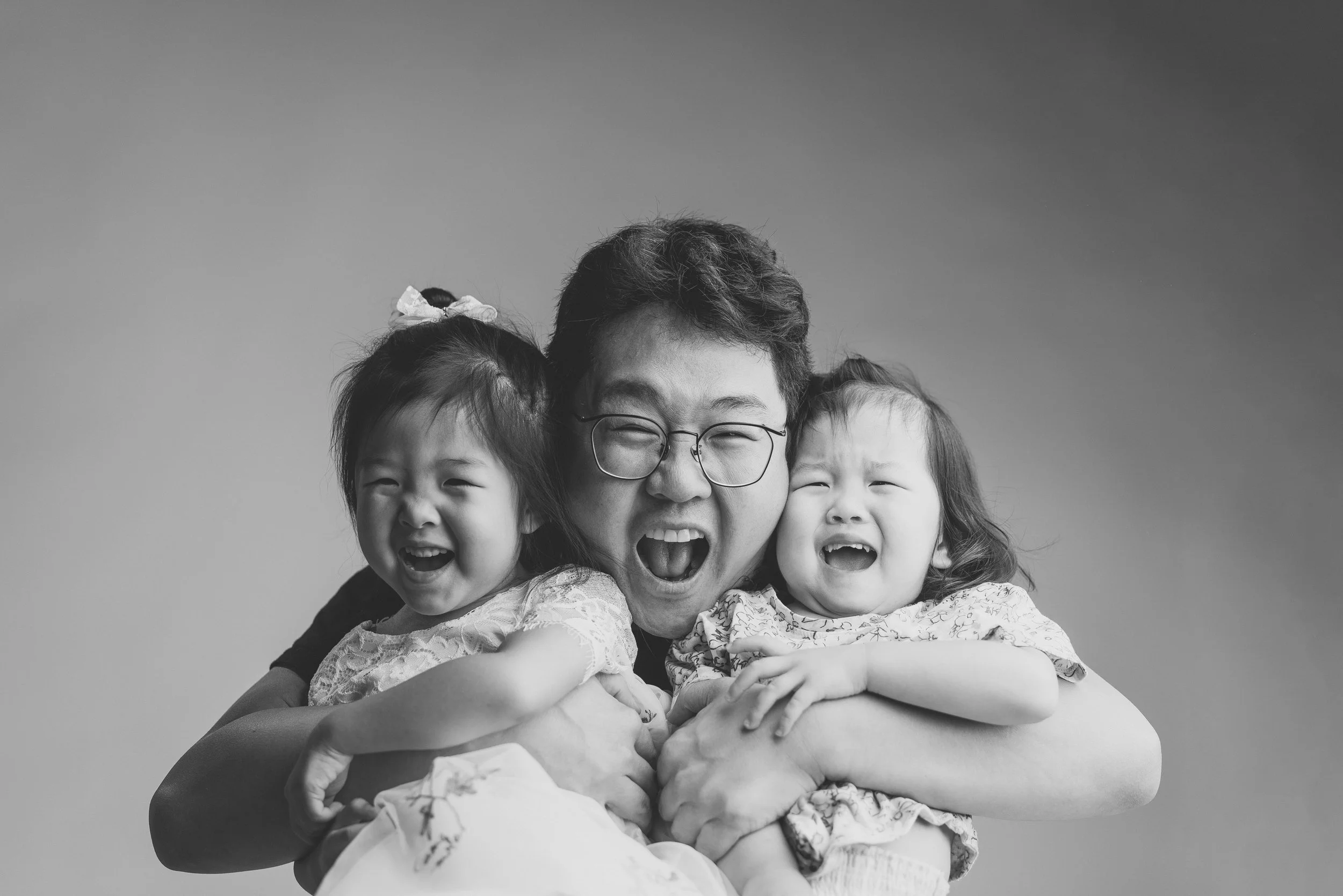 A man with glasses hugging two young girls, all laughing and smiling, in a black-and-white photo.