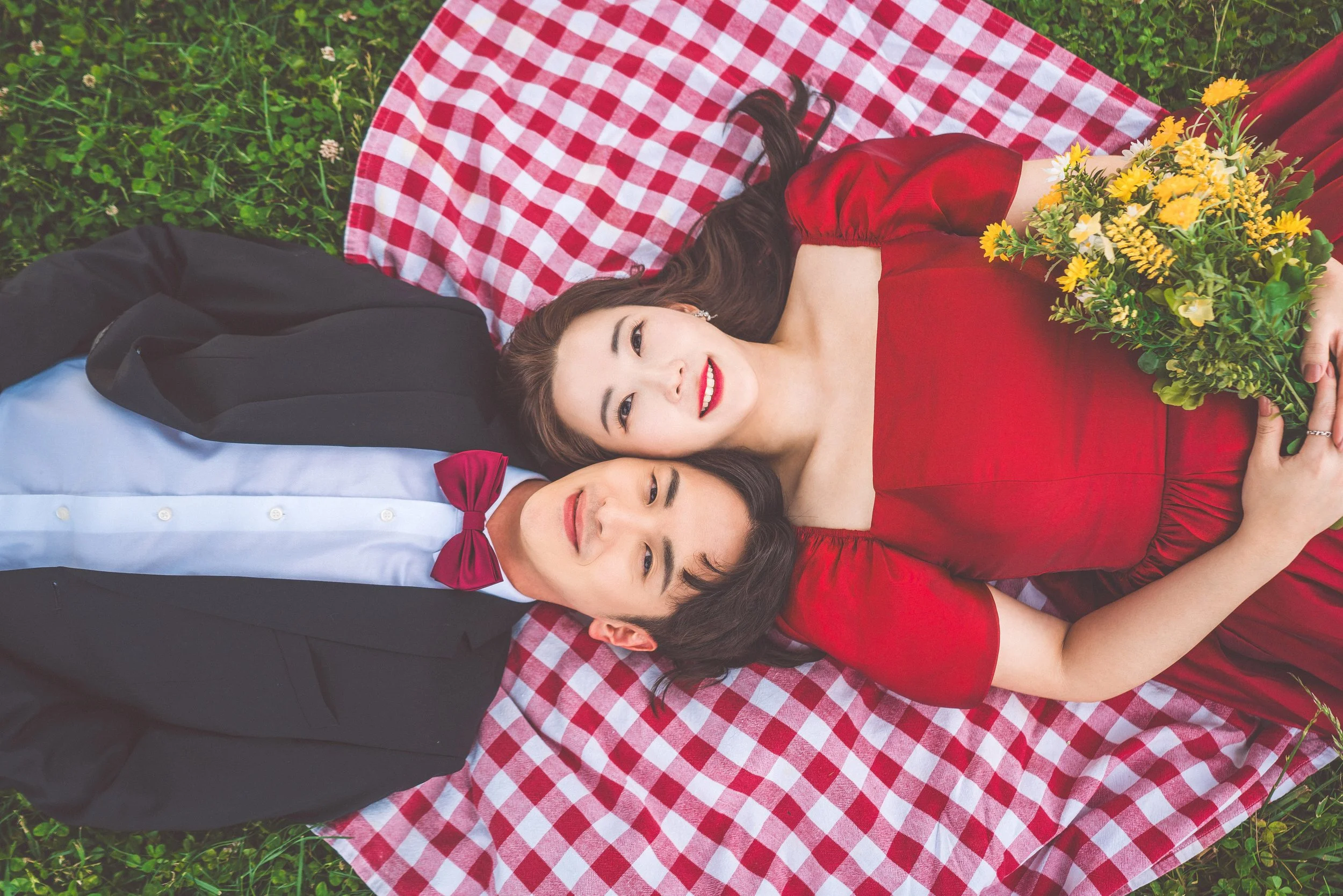 A man and woman lying on a checkered picnic blanket on the grass. The woman is wearing a red dress and holding a bouquet of yellow flowers, while the man is dressed in a black suit with a white shirt and red bow tie. They are looking up and smiling.