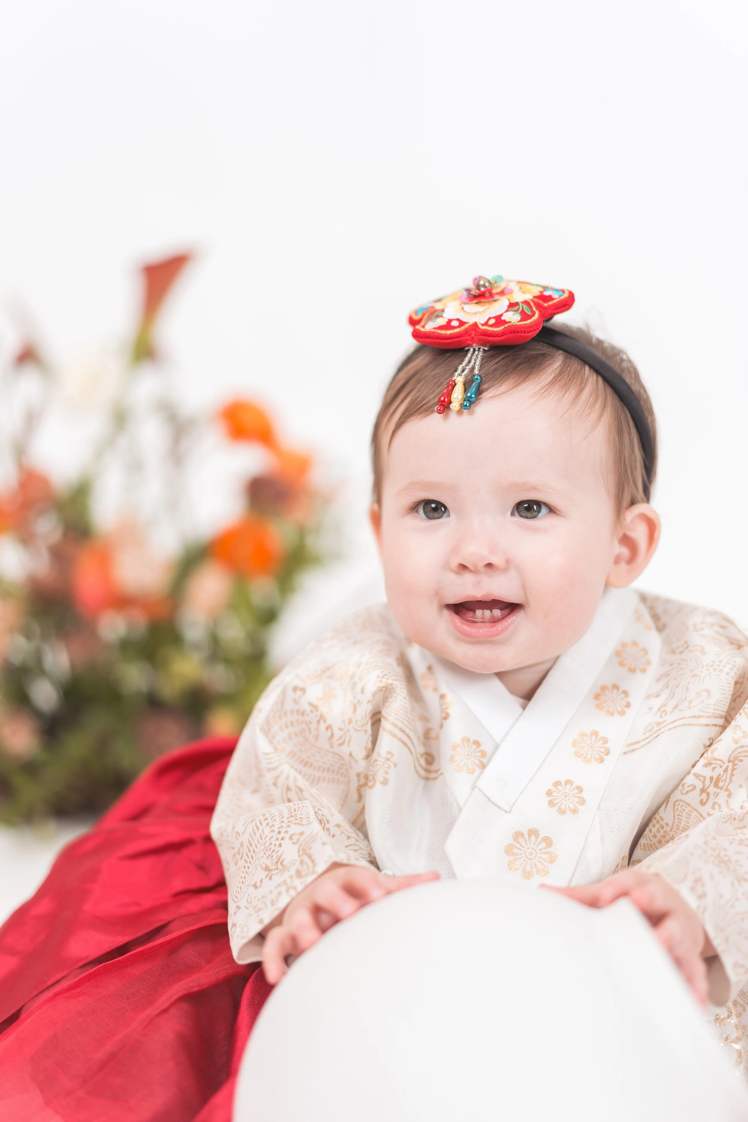 A smiling baby girl wearing traditional Japanese clothing and a decorative headband, sitting with a white balloon, with blurred colorful flowers in the background.