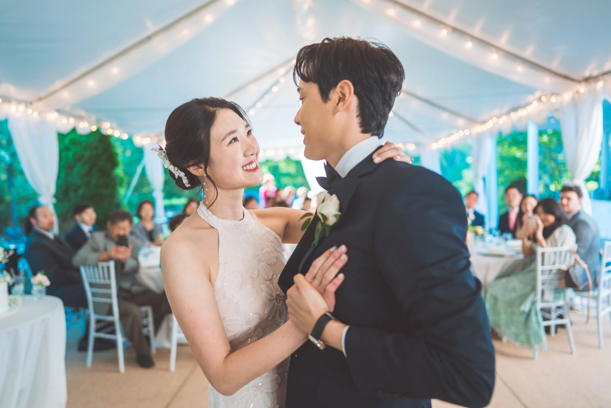 A bride and groom dancing at their wedding reception under a decorated tent, with seated guests in the background.