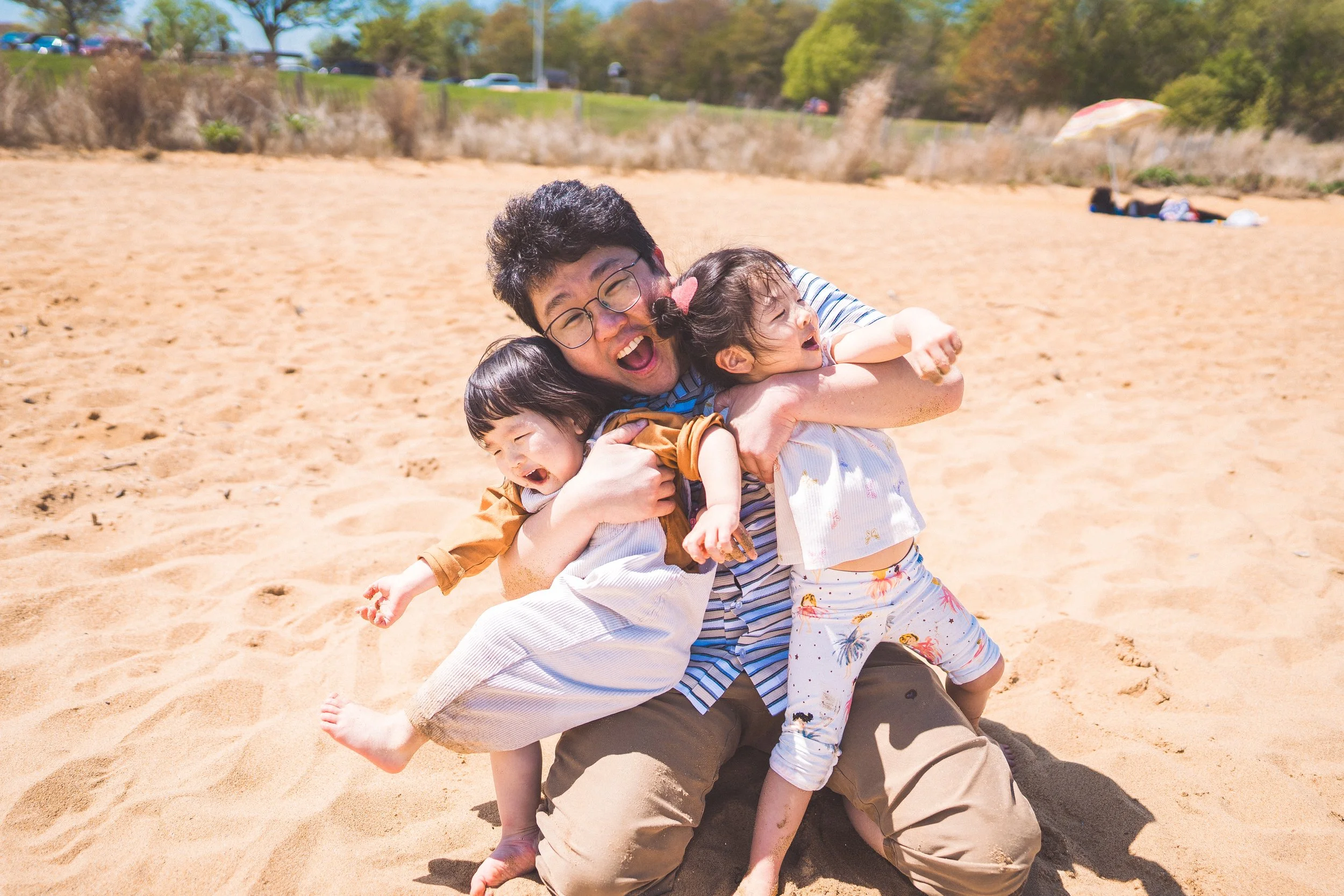 A man playing with two young children on a sandy beach, happily hugging and laughing.