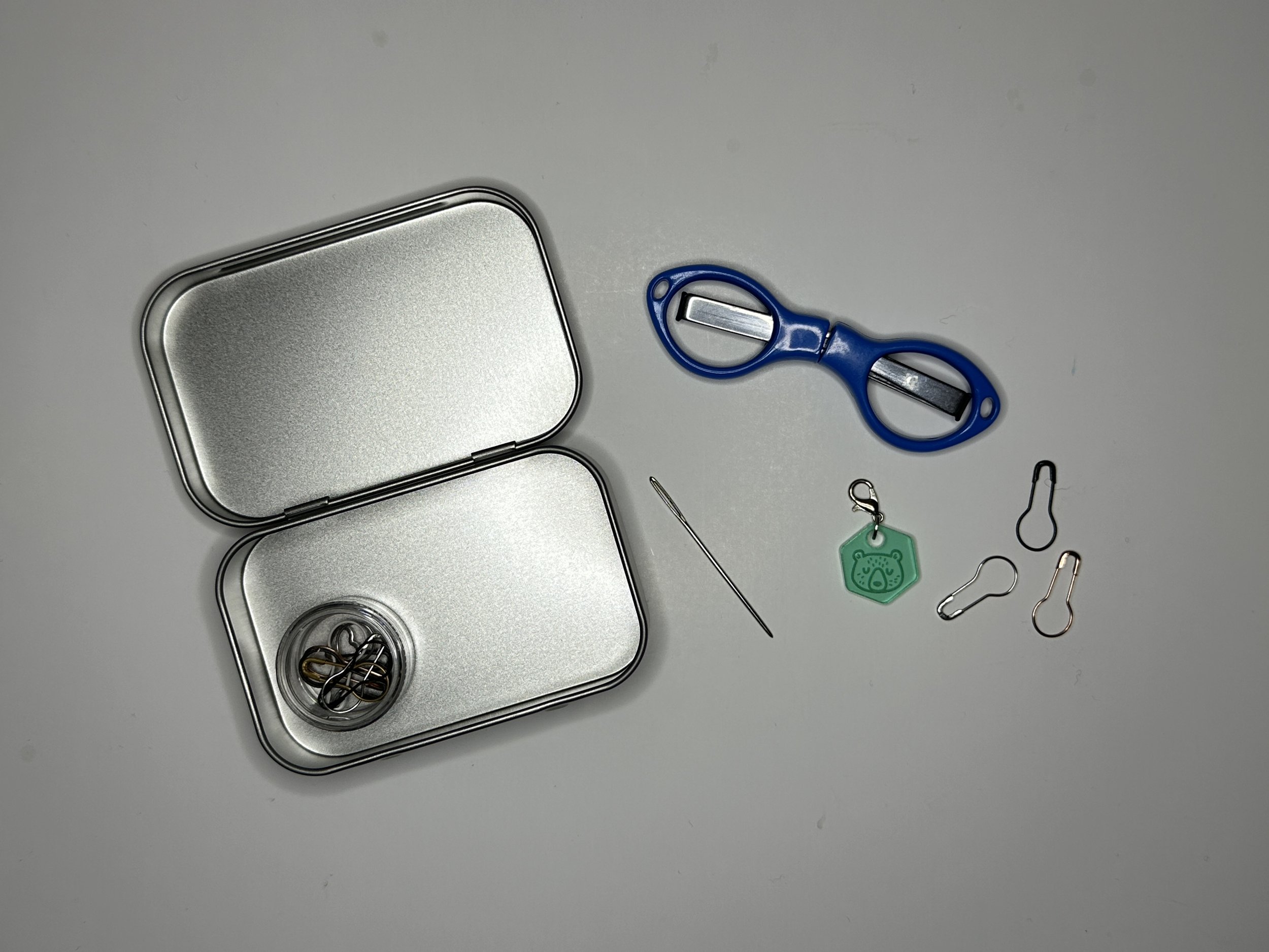 Small tin with a container of safety stitch markers in side. on the table rests foldable scissors, stitch markers, a progress keeper, and darning needle