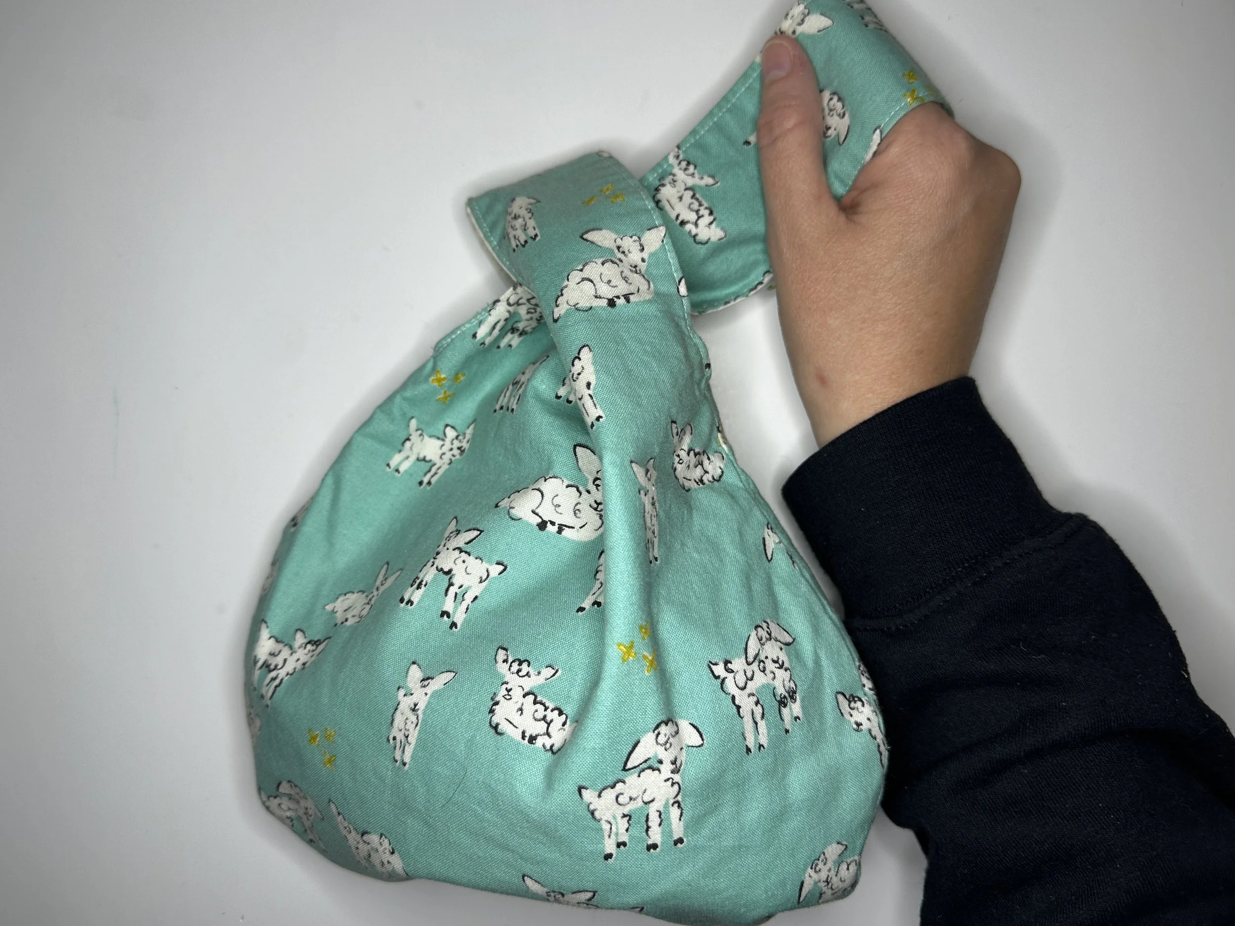 Hand with a black sleeve shirt holding a green small project bag with lambs on it