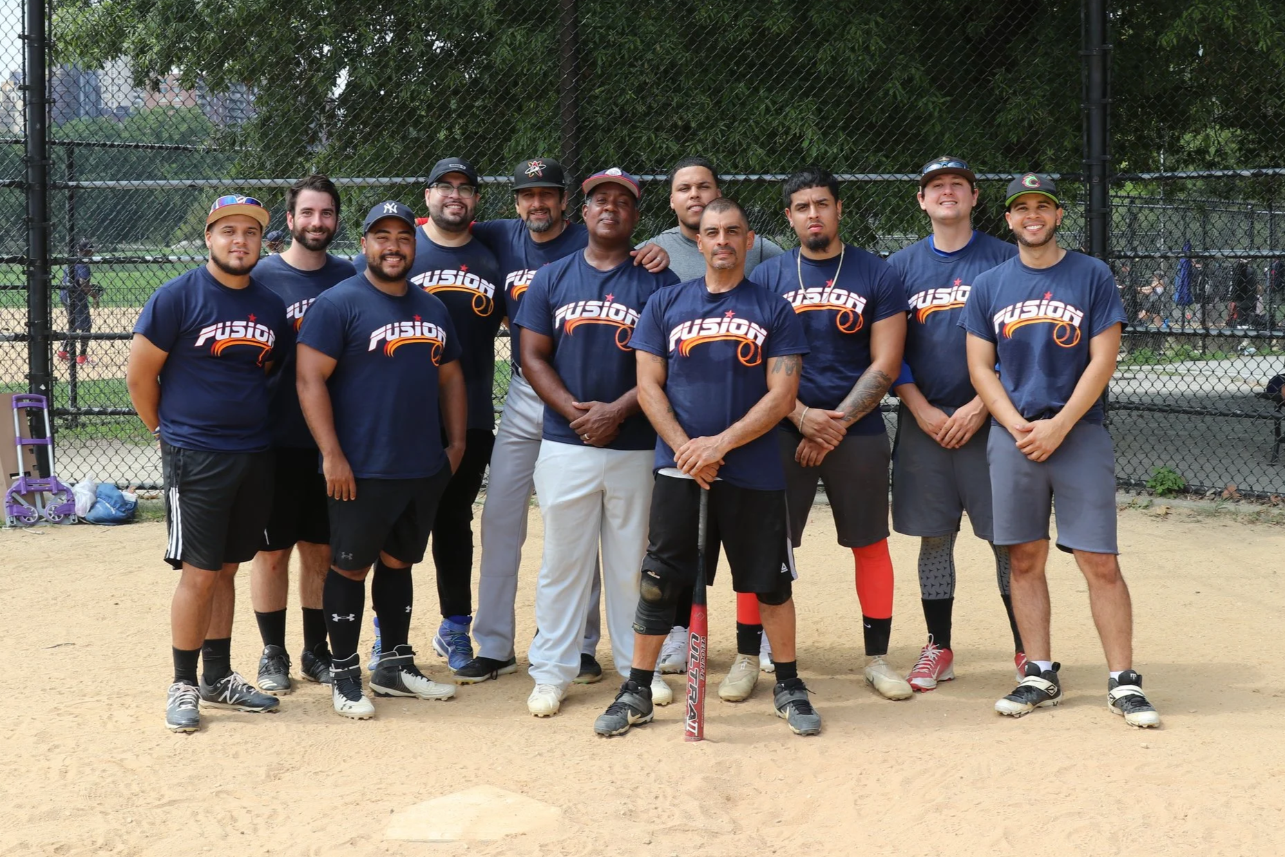 A group of people wearing matching blue shirts with a logo, standing on a baseball field in front of a chain-link fence.