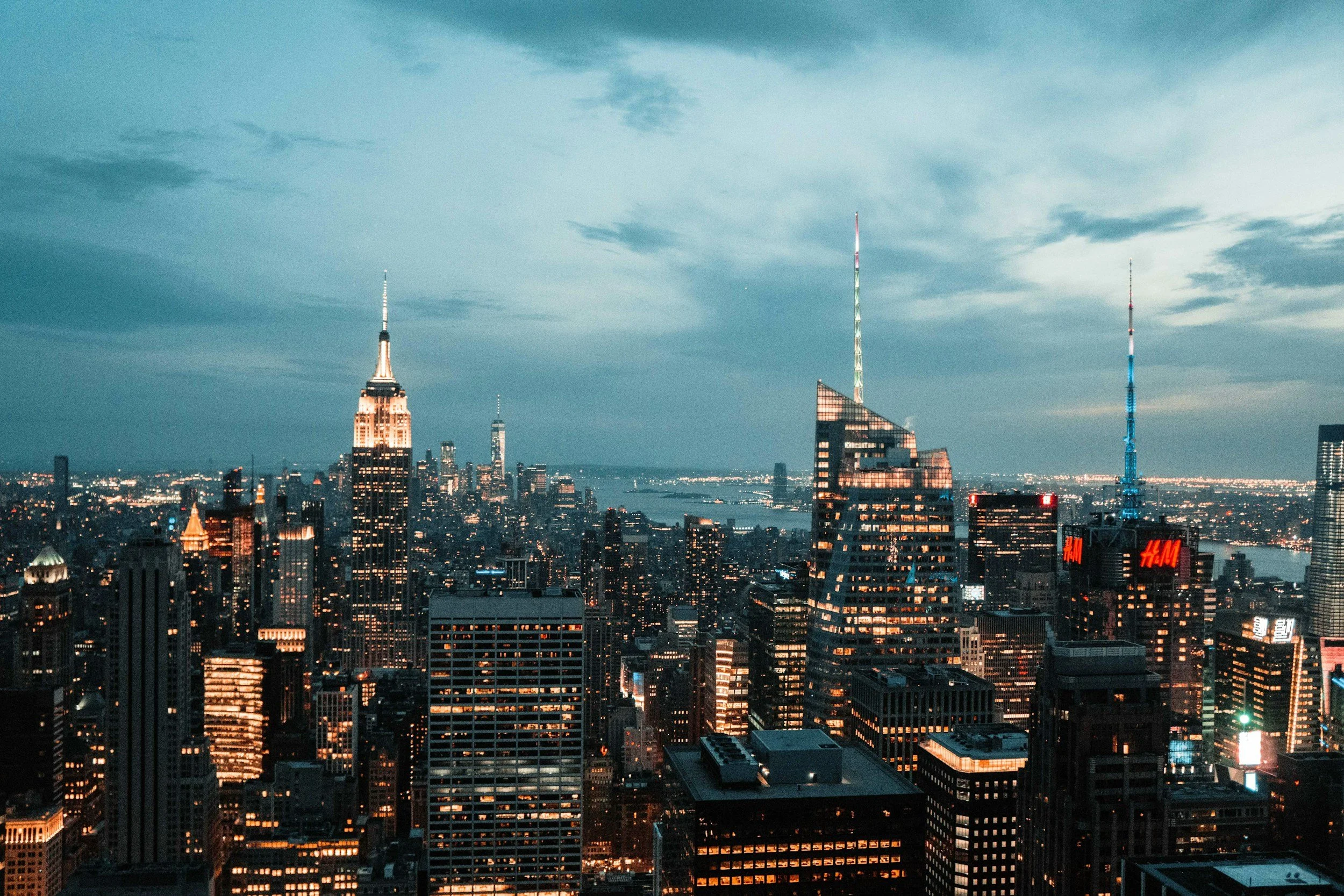Nighttime view of New York City skyline with illuminated skyscrapers, including the Empire State Building and One World Trade Center, under a cloudy sky.