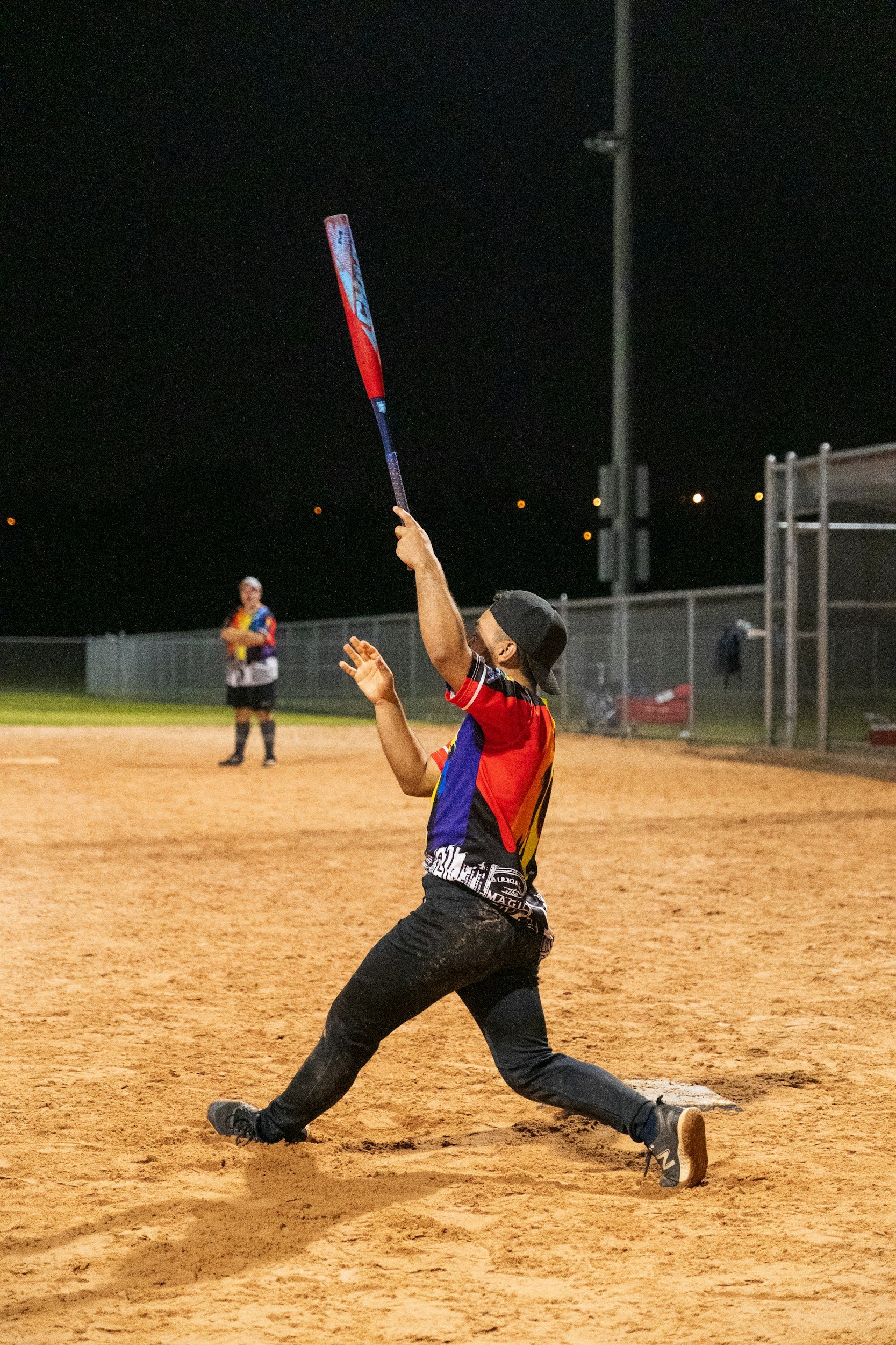 A baseball player in a colorful jersey and black pants is mid-swing at night on a dirt field. He is holding a baseball bat, looking upward, with another player in the background.