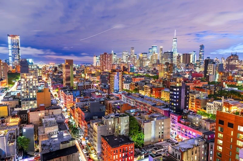 Cityscape of New York City skyline at dusk with illuminated buildings and a partly cloudy sky.