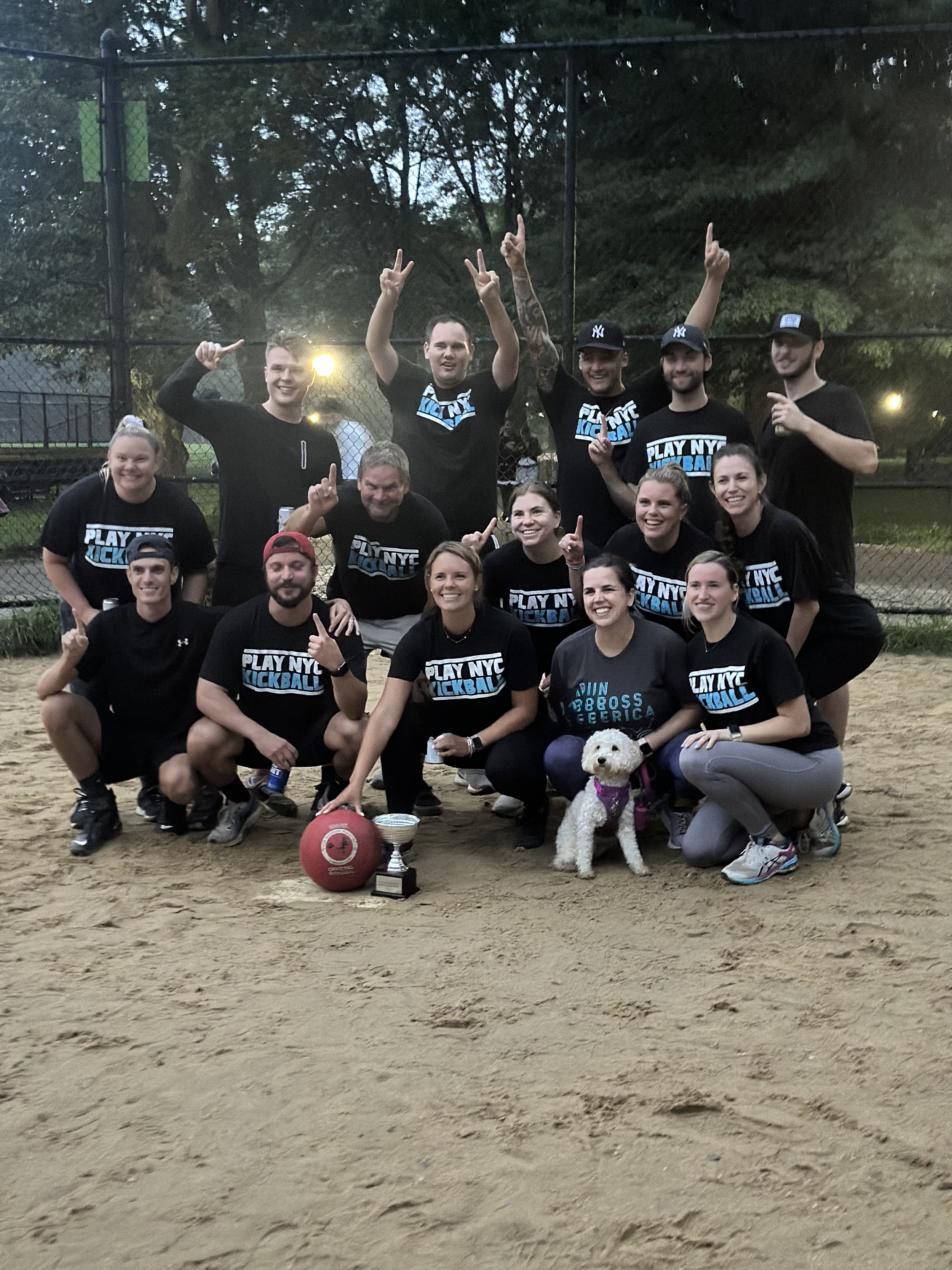 A group of people, some wearing matching black t-shirts that say 'PLAY NYC KICKBALL,' are gathered on a dirt field, celebrating a victory with some holding up peace signs and others smiling. In front, a small white dog is sitting next to a red kickball, a trophy, and a small bowl on the ground. The background shows a fence and trees.