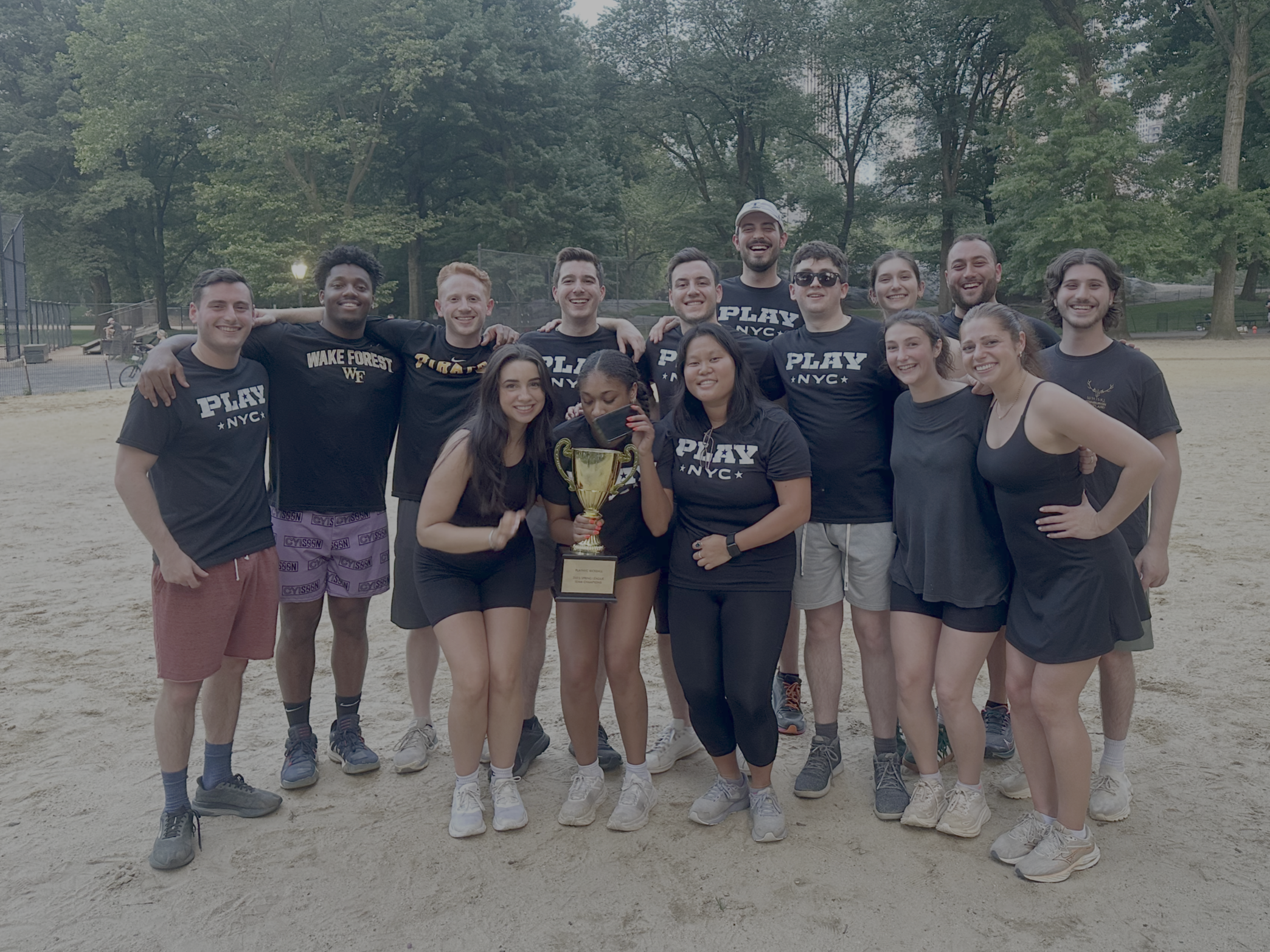 A group of young people standing together on a sandy sports field, holding a trophy, celebrating after a sports event, with trees and a park in the background.