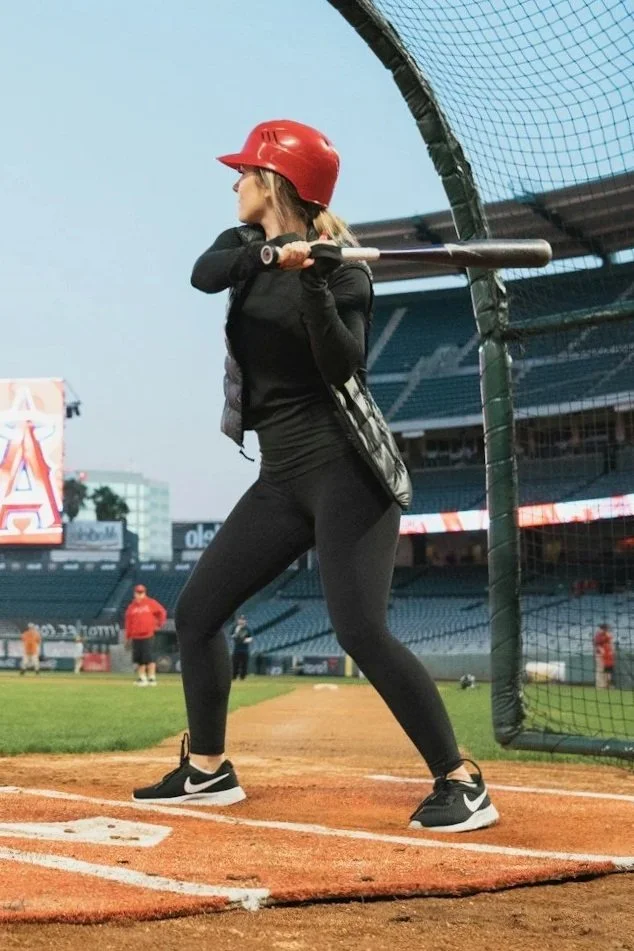 A woman in black athletic clothes and a red helmet holding a baseball bat, standing in a batting cage at a baseball stadium.