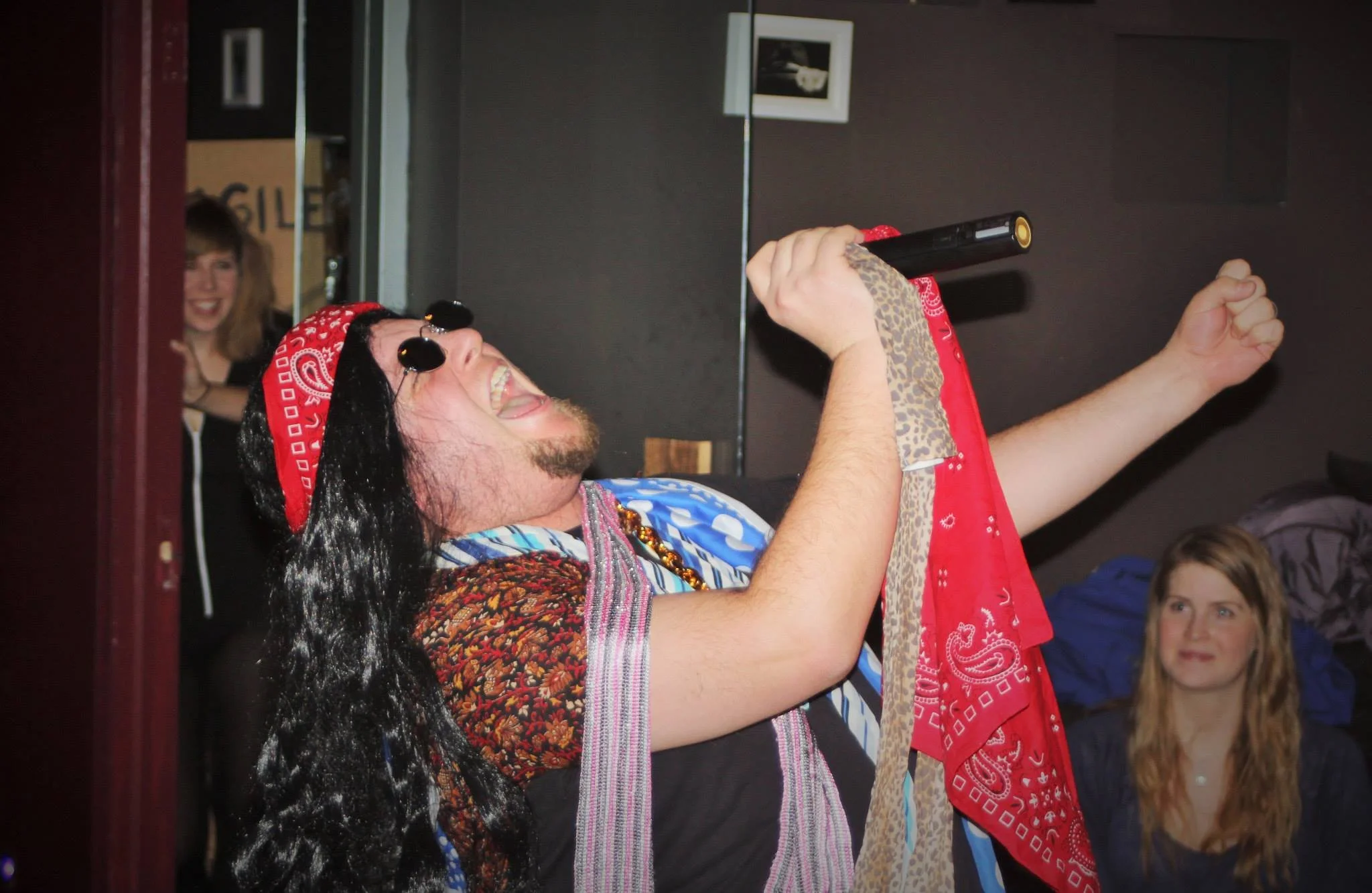 A man dressed in a colorful shirt and sunglasses, singing with a microphone, wearing a red bandana, in a room with two women watching and smiling.