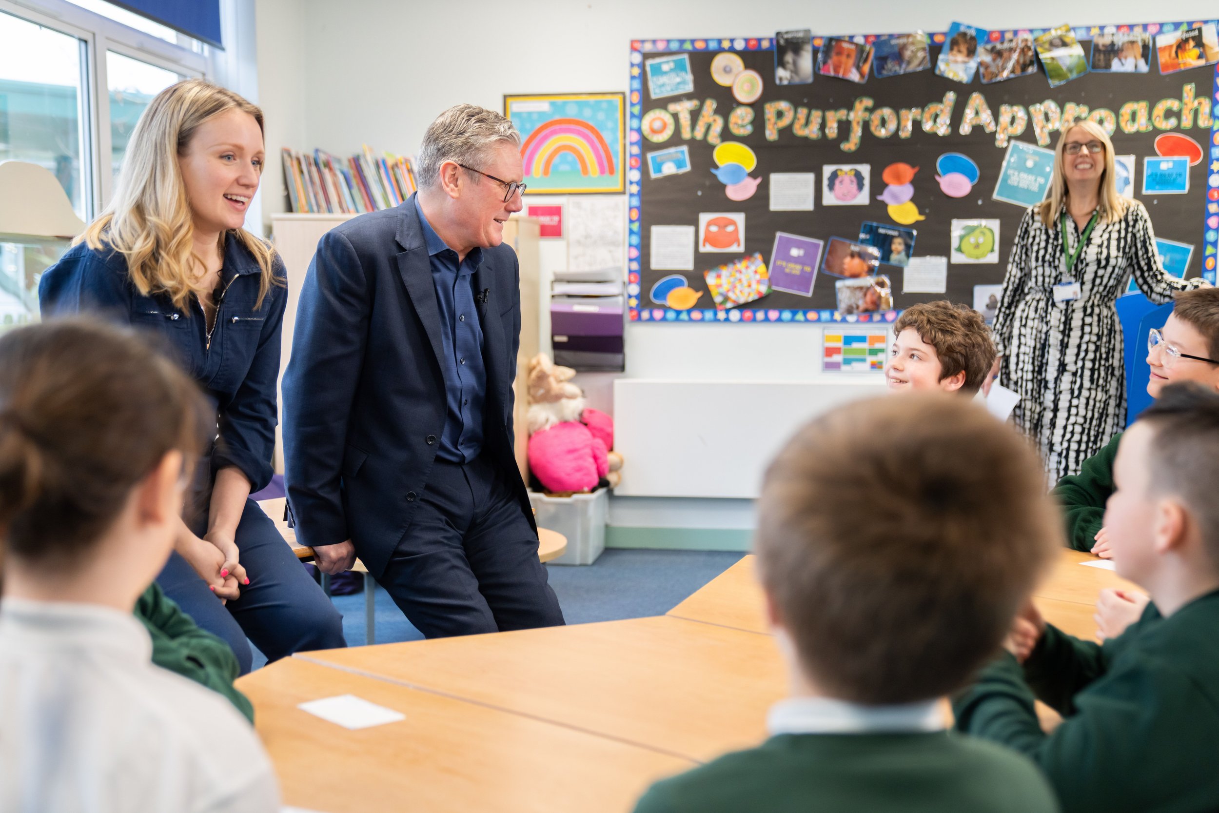 A classroom scene with Emily and Keir Starme smiling at students seated at desks. The background features a bulletin board with colorful decorations