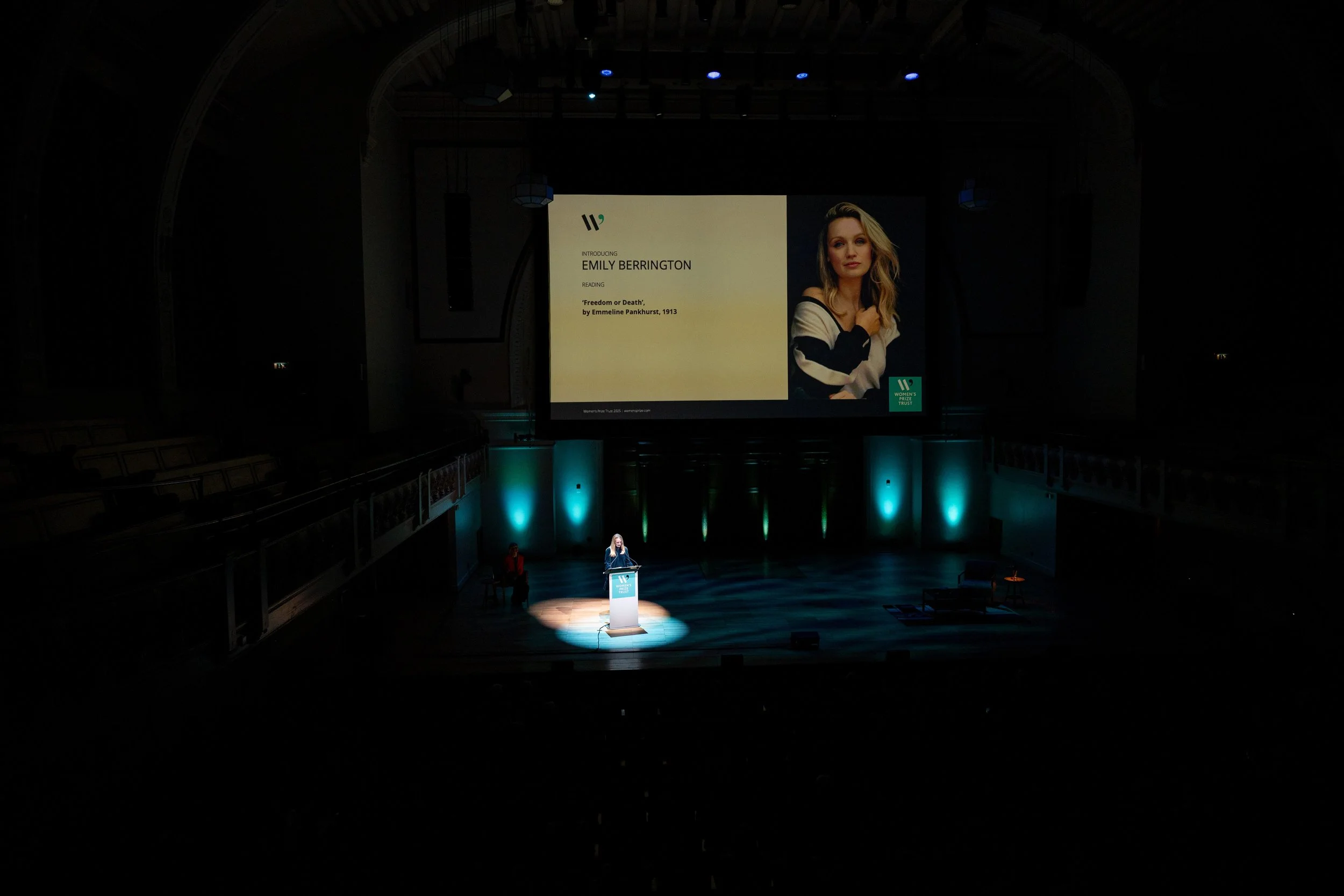 A woman is speaking at a podium on a stage illuminated with spotlight, with a large screen behind her displaying her name, Emily Berrington, and a quote from a reading. The background features teal-colored lighting accents, and the auditorium appears dark with empty seats visible to the sides.