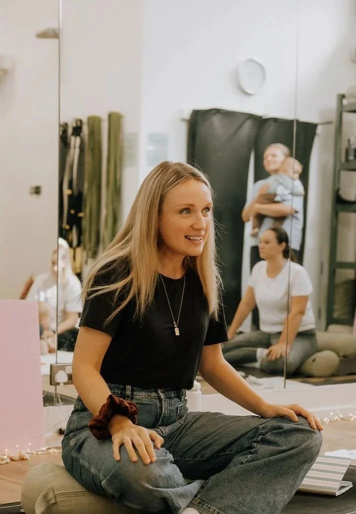 A woman sitting cross-legged on a cushion, smiling, in a room with mirrors and other people in the background.