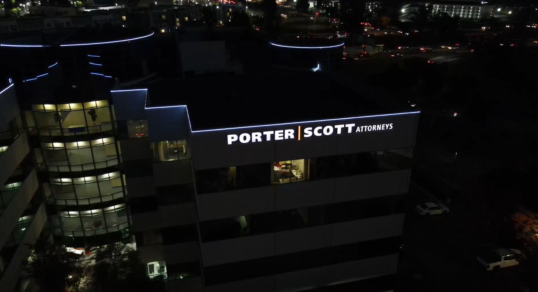 Nighttime aerial view of a multi-story building with illuminated signage reading 'PORTER | SCOTT ATTORNEYS' on the top level, with office lights visible through the windows.